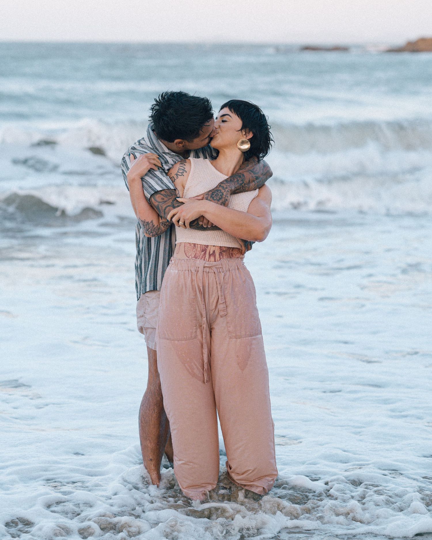 A tattooed couple kisses and embraces while standing in the shallow ocean surf, the woman wearing blush linen pants.