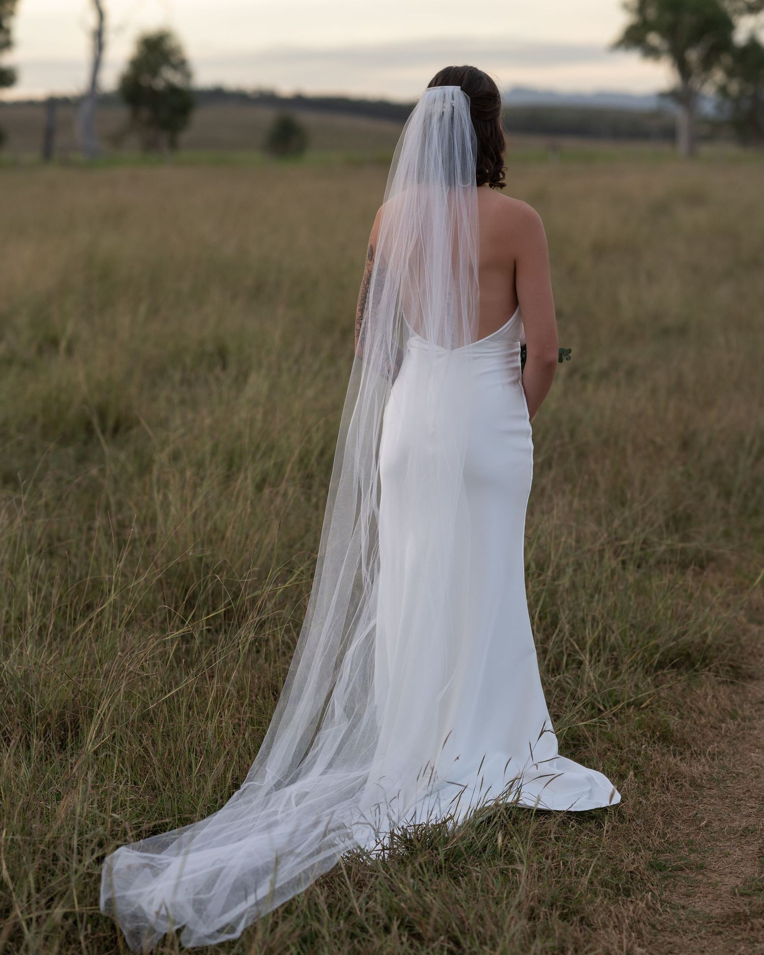 Bride in a fitted white gown and long sheer veil stands facing away in a grassy field at dusk.