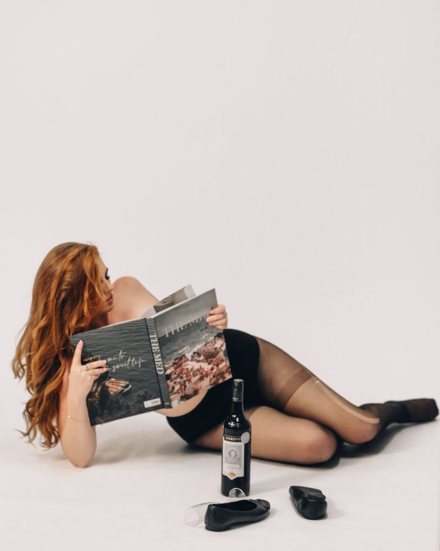 Woman with long red hair lies on a white studio floor reading a book beside a wine bottle and black shoes.