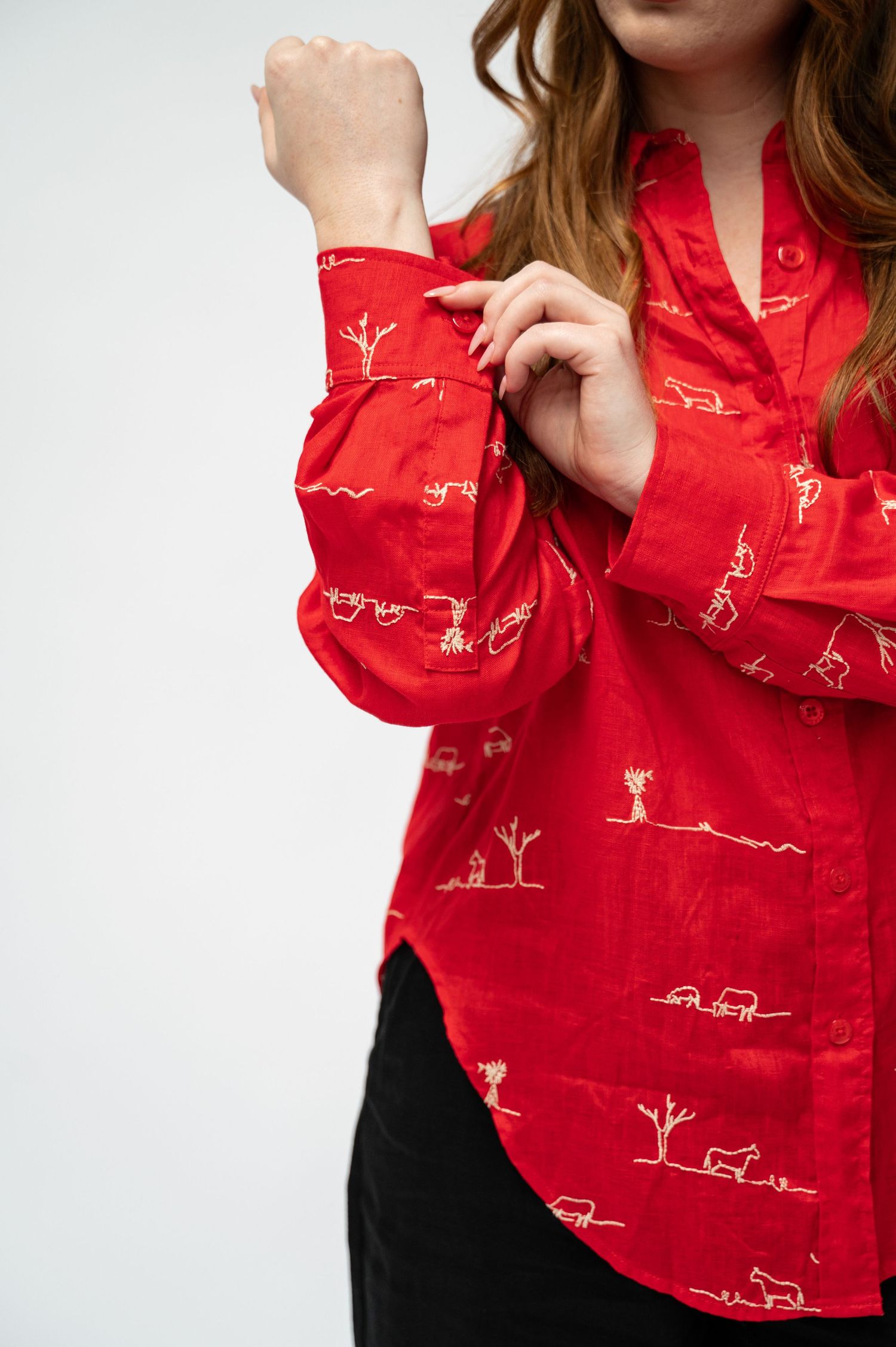 Close-up of a woman adjusting the cuff of a red button-up shirt printed with cream line drawings of trees and animals.