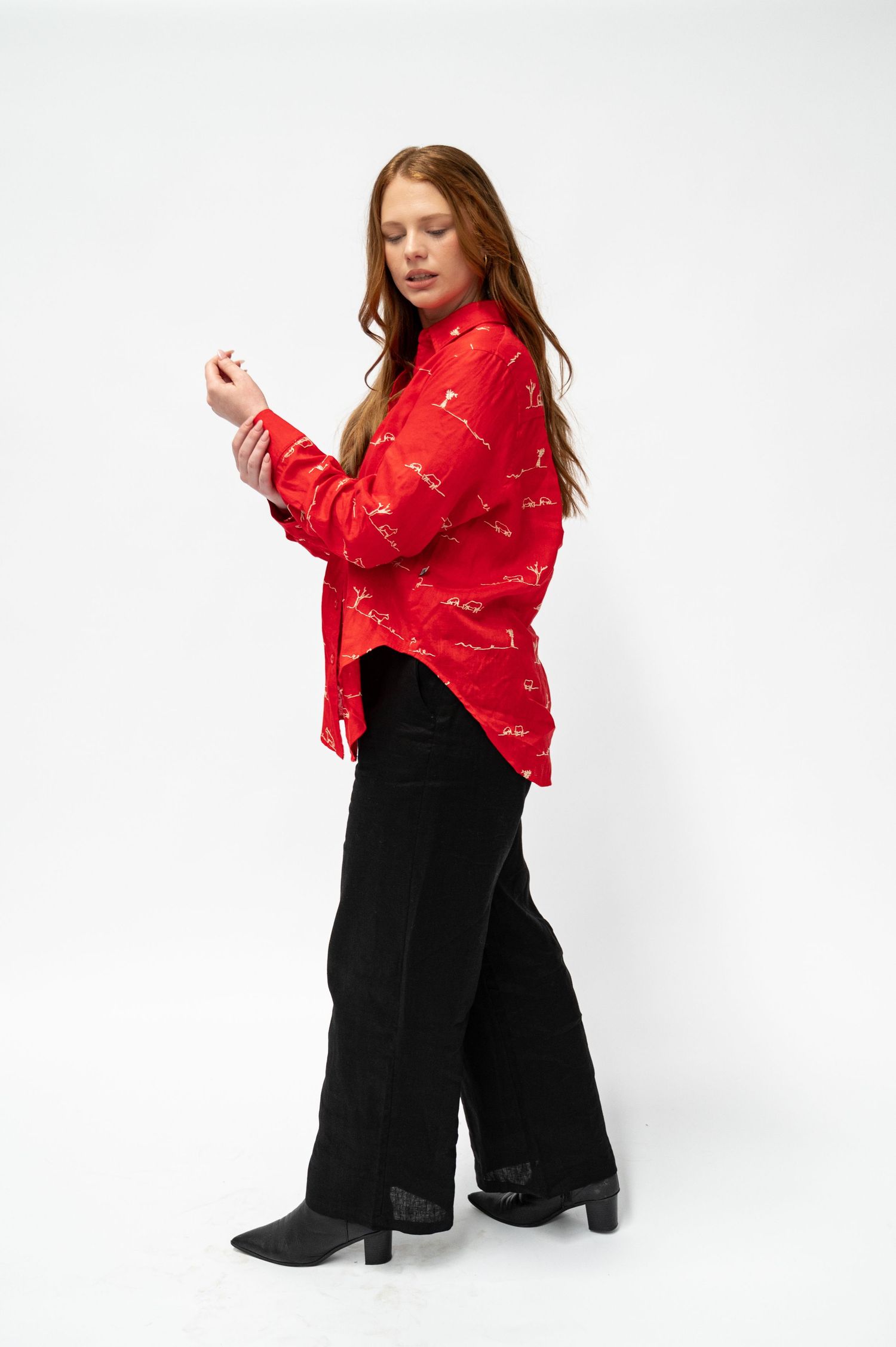 A woman models a red long-sleeve linen shirt with a white graphic print, black wide-leg corduroy pants and black heeled ankle boots.