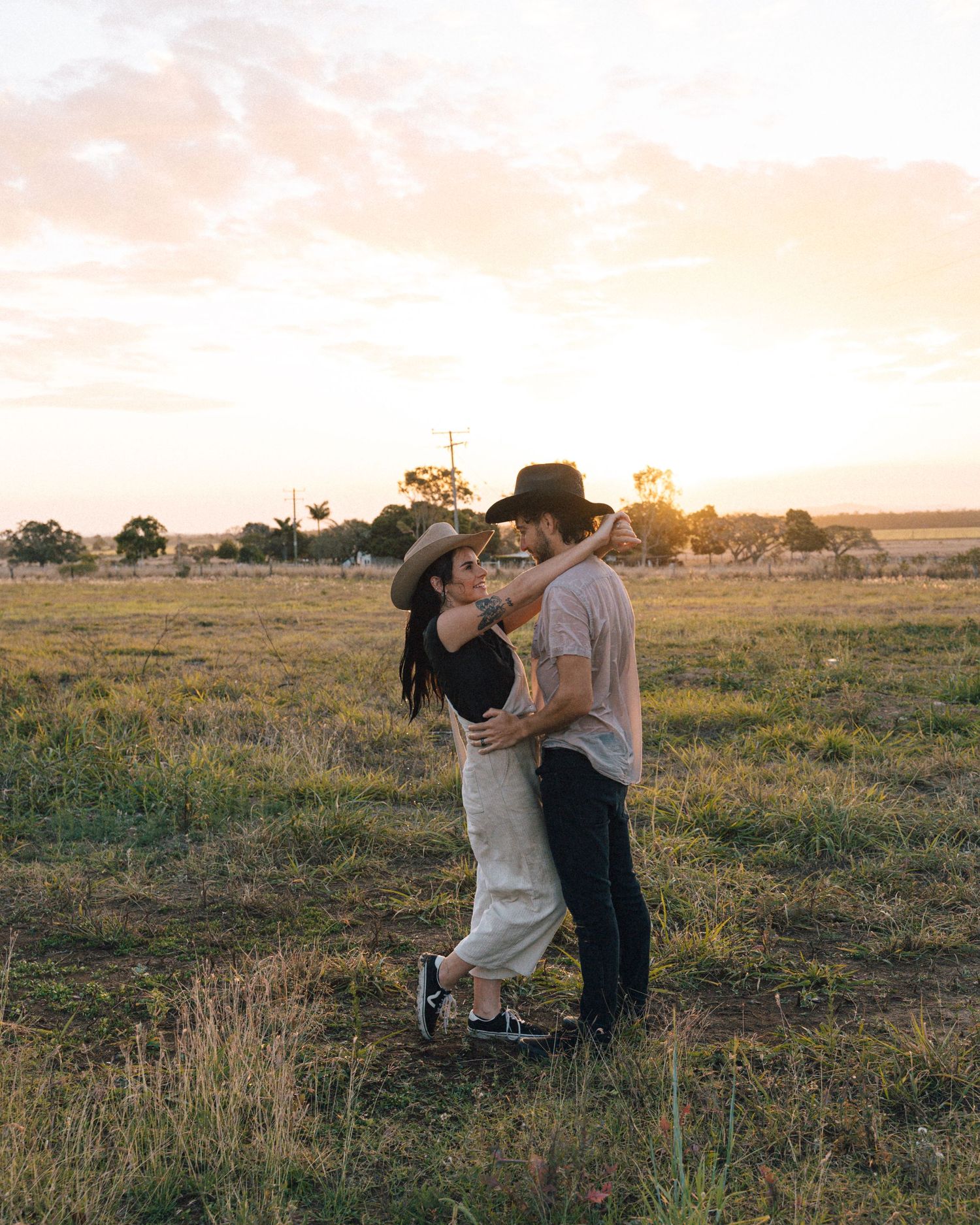 Couple in cowboy hats embrace in a wide open grassy field beneath a soft pastel sunset sky.