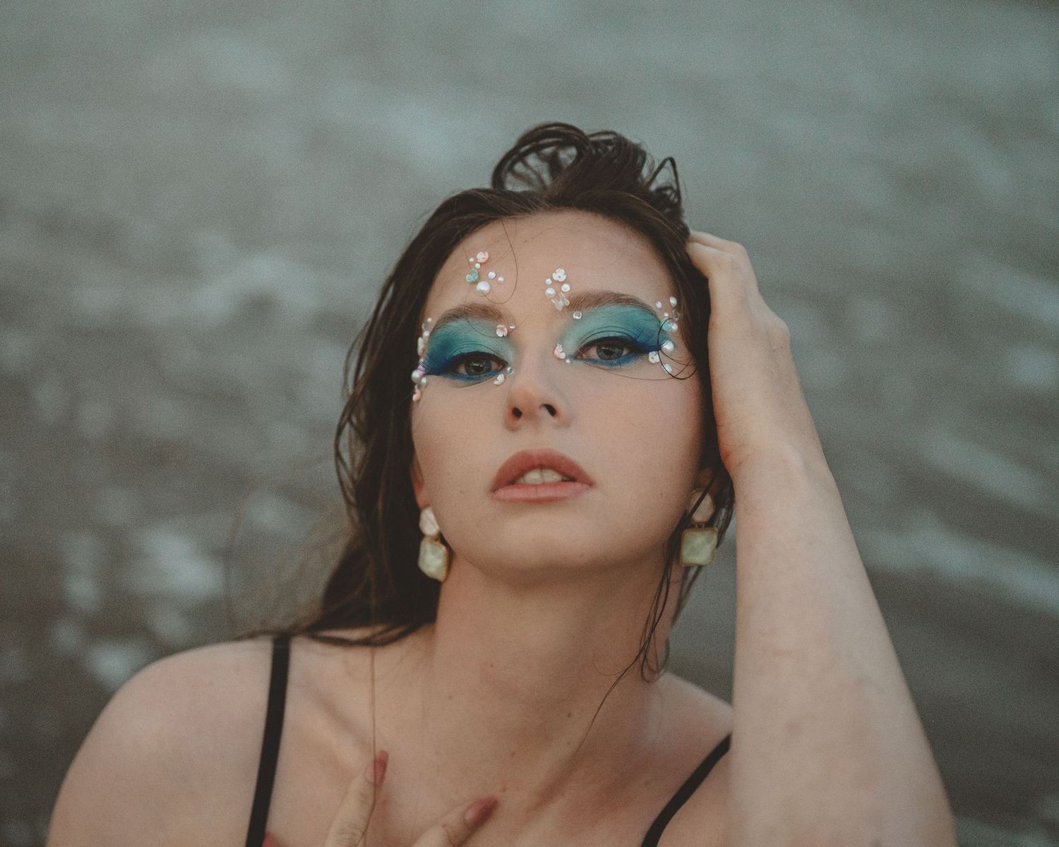 Beach portrait of a woman with wet hair, blue eyeshadow and gem face jewels, wearing drop earrings with moody ocean tones behind her.