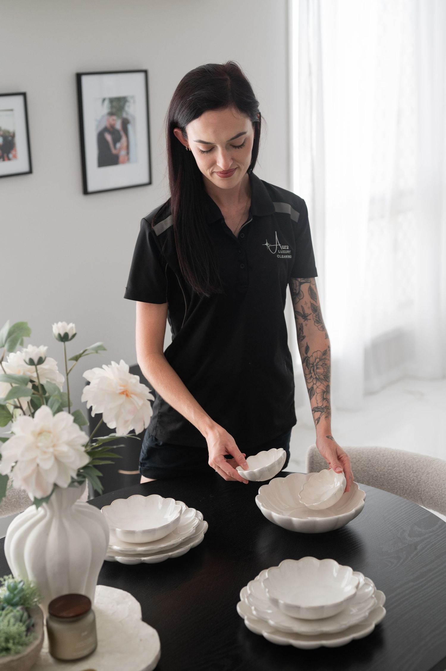 Woman in black uniform with tattoos styling white scalloped bowls and plates on a black dining table with white flowers.