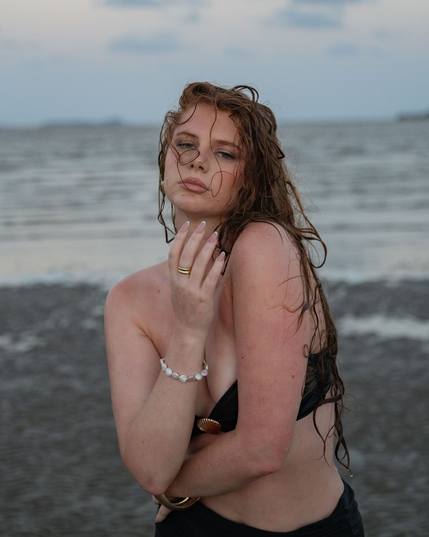 Woman with long wet auburn hair stands at the shoreline wearing a pearl bracelet and gold ring under a cloudy sky.