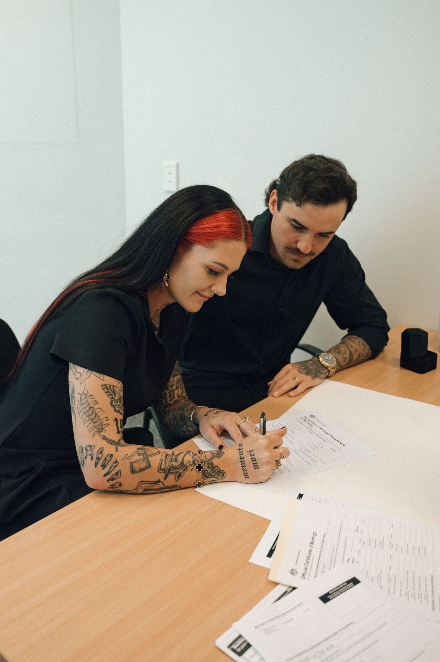 Smiling couple in black outfits with tattoos sign marriage documents together at a wooden table.