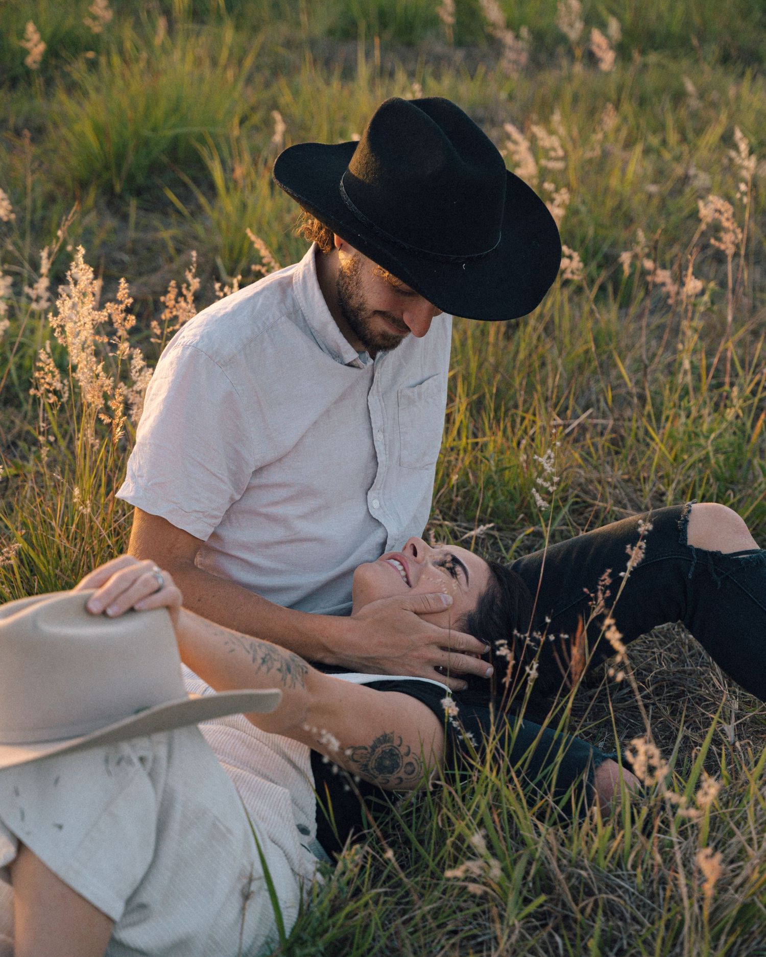 A man in a black cowboy hat sits in tall grass at sunset, gently holding a smiling woman lying across his lap.