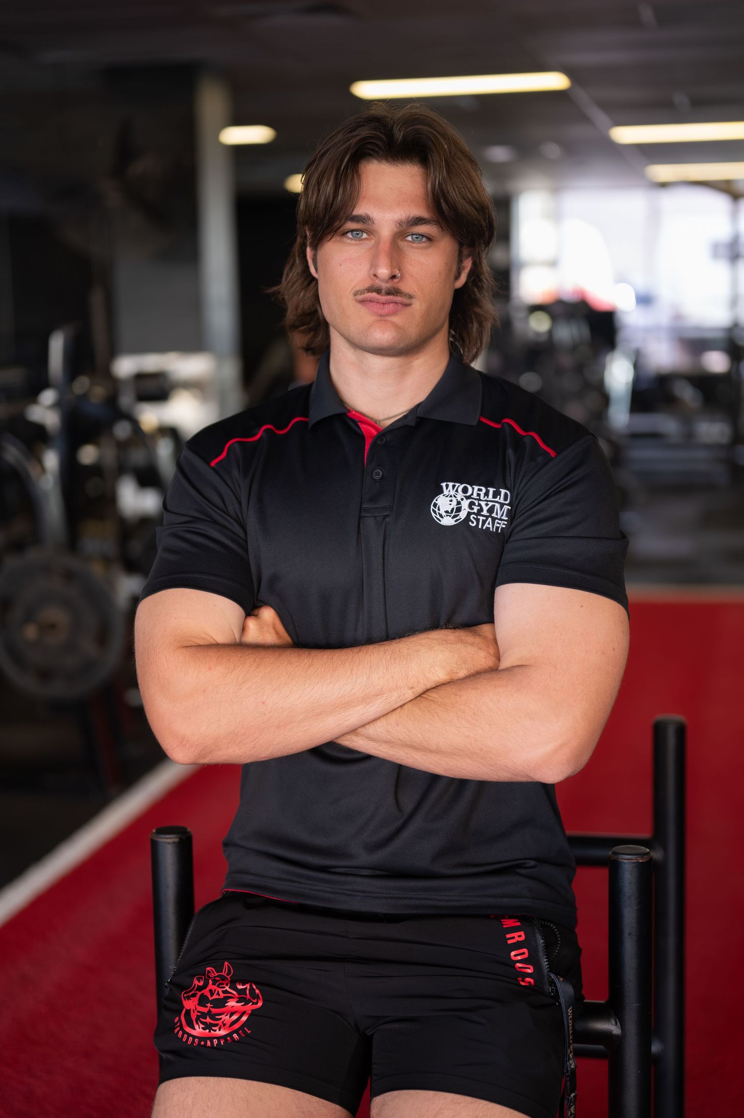 Young man with shoulder-length brown hair and a mustache poses with arms crossed in a gym wearing a World Gym staff shirt.