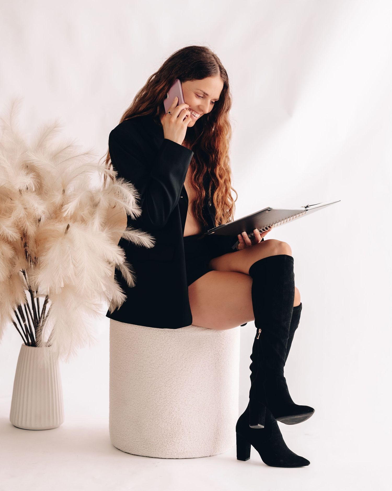 Woman in a black blazer and suede boots sits on a white pouf, talking on her phone while holding a notebook beside pampas grass.