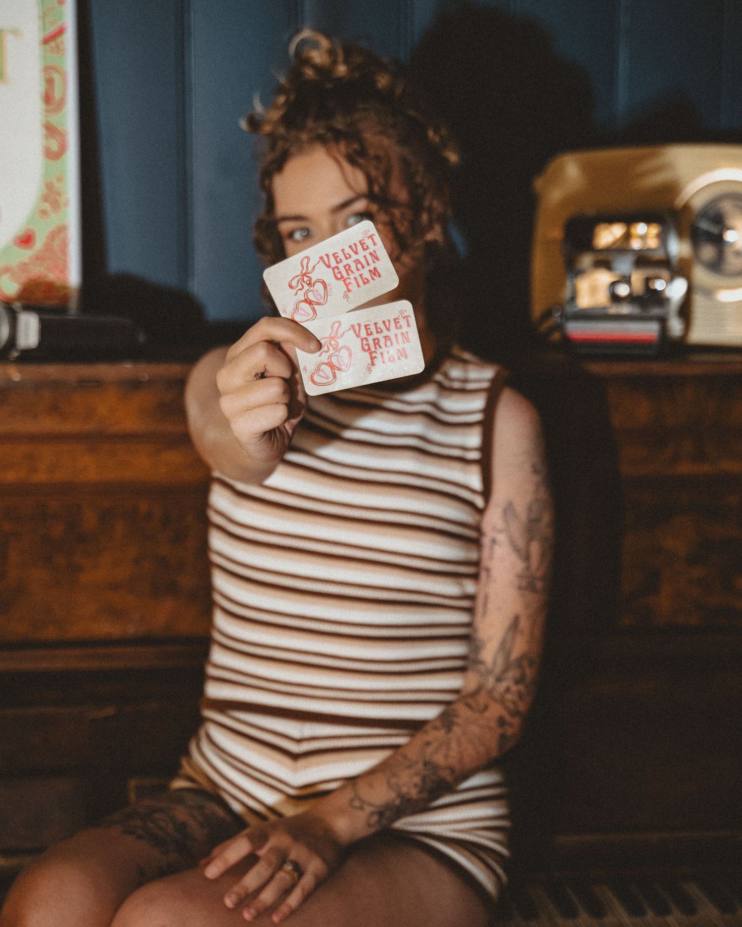 Woman in a brown striped sleeveless top with tattoos holds up two Velvet Grain Film cards in front of her face beside a vintage camera.