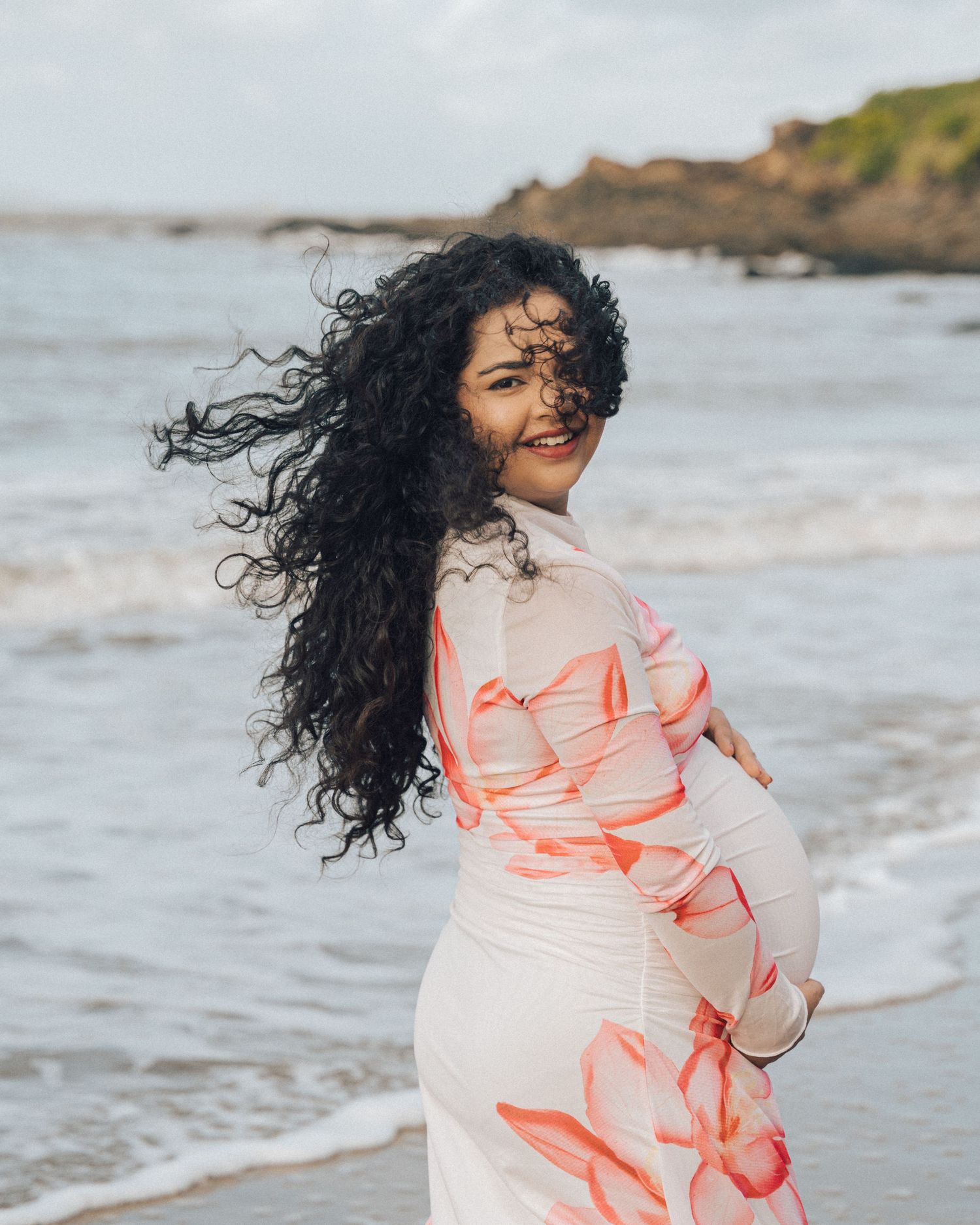 Smiling pregnant woman with long curly black hair in a pink floral maxi dress stands on a windswept beach by the ocean.