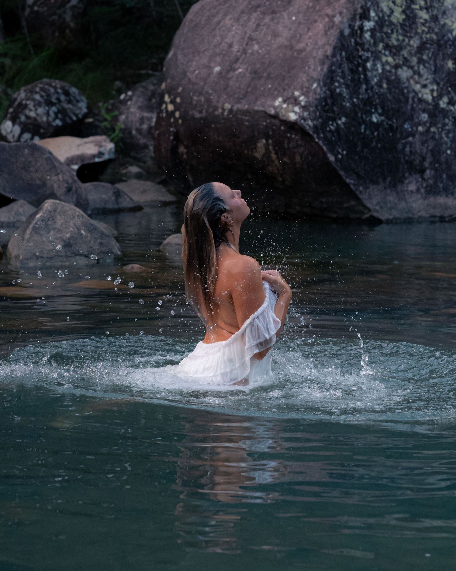 Woman in a white wrap rises from a calm rocky lake with water splashing around her, large boulders and trees behind.