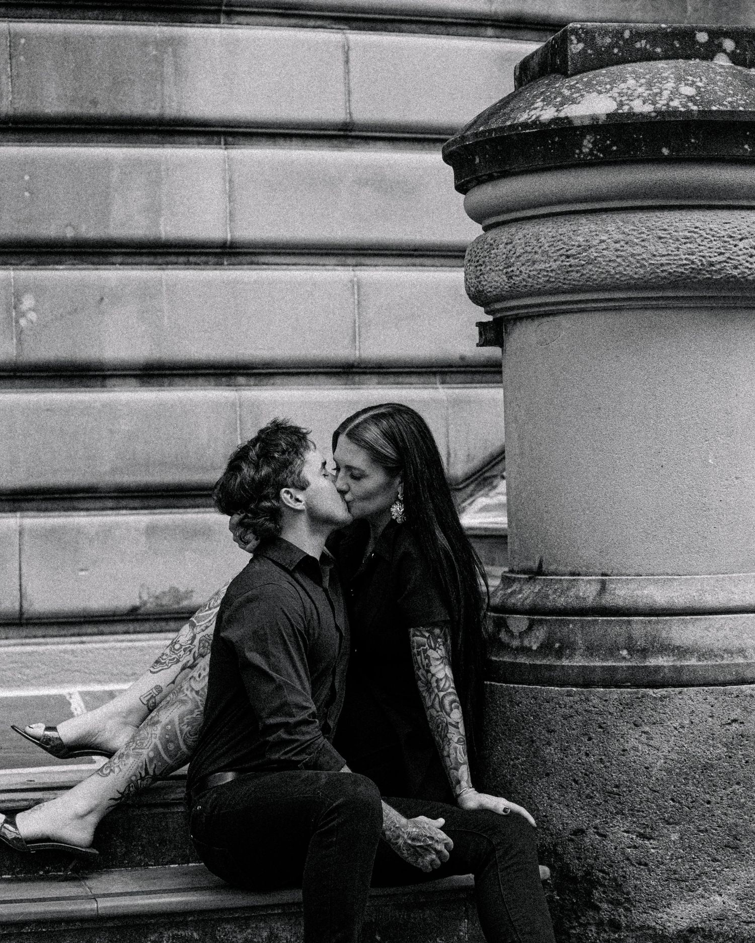 Black and white photo of a tattooed couple kissing while seated on stone steps beside a large rounded stone pillar.
