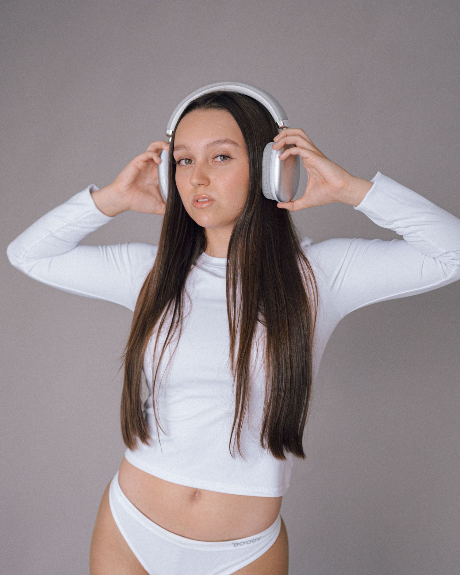 Woman with long brown hair wearing white over-ear headphones and a white long-sleeve crop top, posing on a gray background.