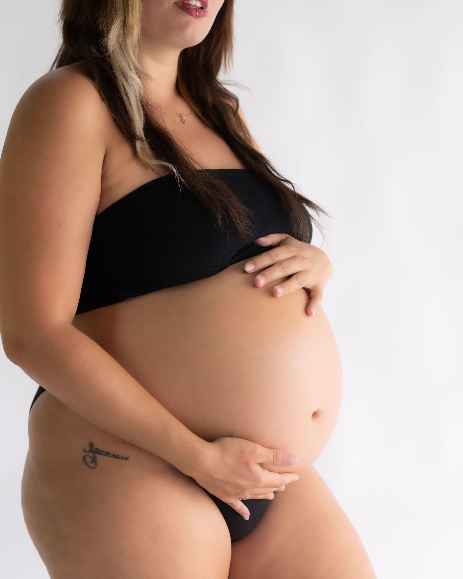 Pregnant woman in a black two-piece set cradles her bare belly against a soft white studio background in a maternity portrait.