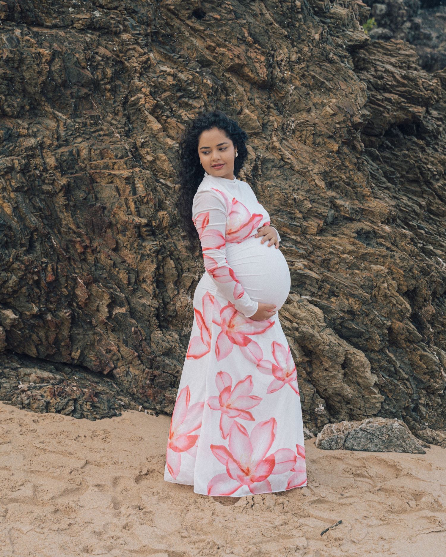 Expectant mother in a long pink floral maxi dress cradles her baby bump on a sandy beach in front of rocky cliffs.