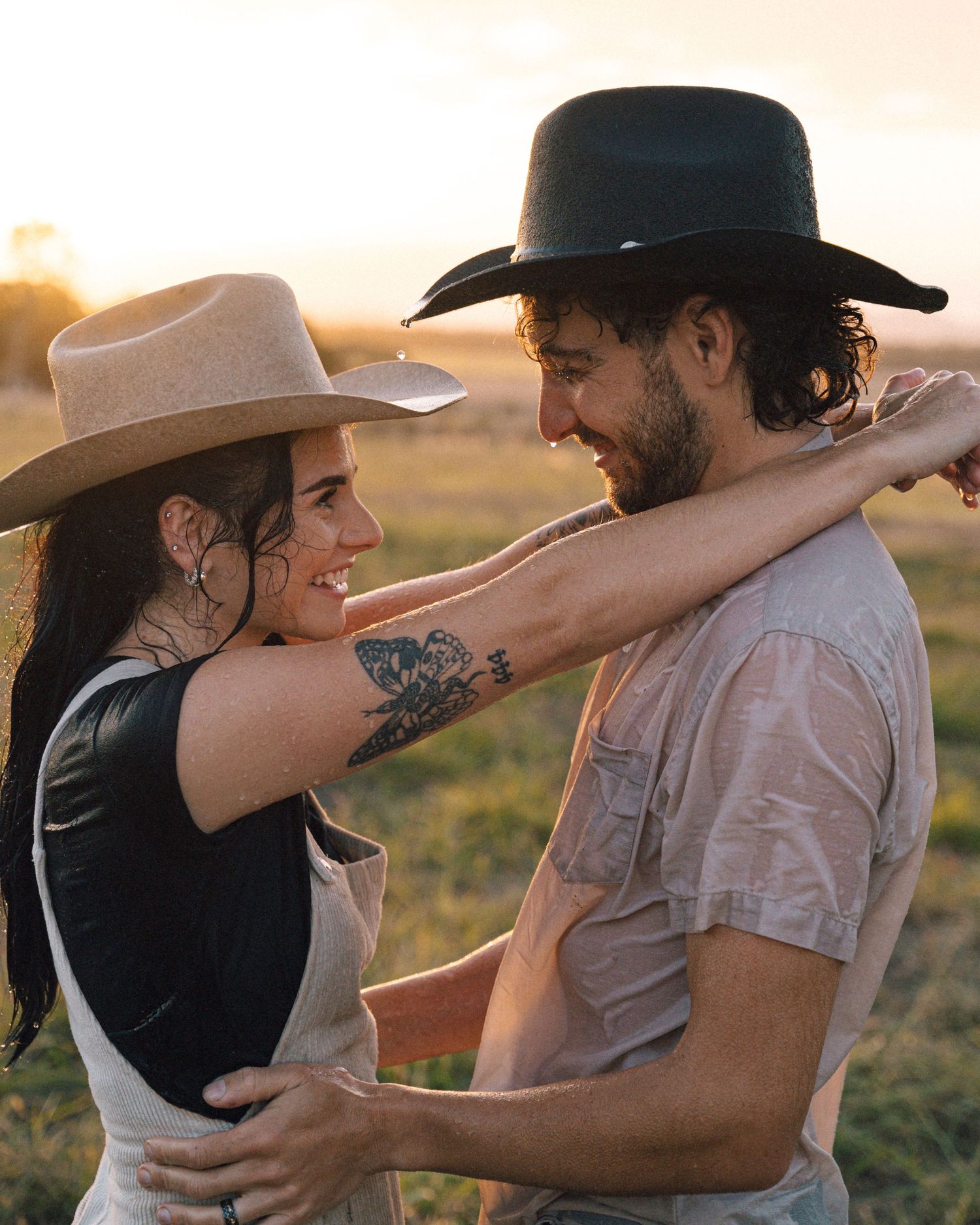 Joyful couple in cowboy hats embracing in a golden field at sunset, the woman showing a floral arm tattoo.