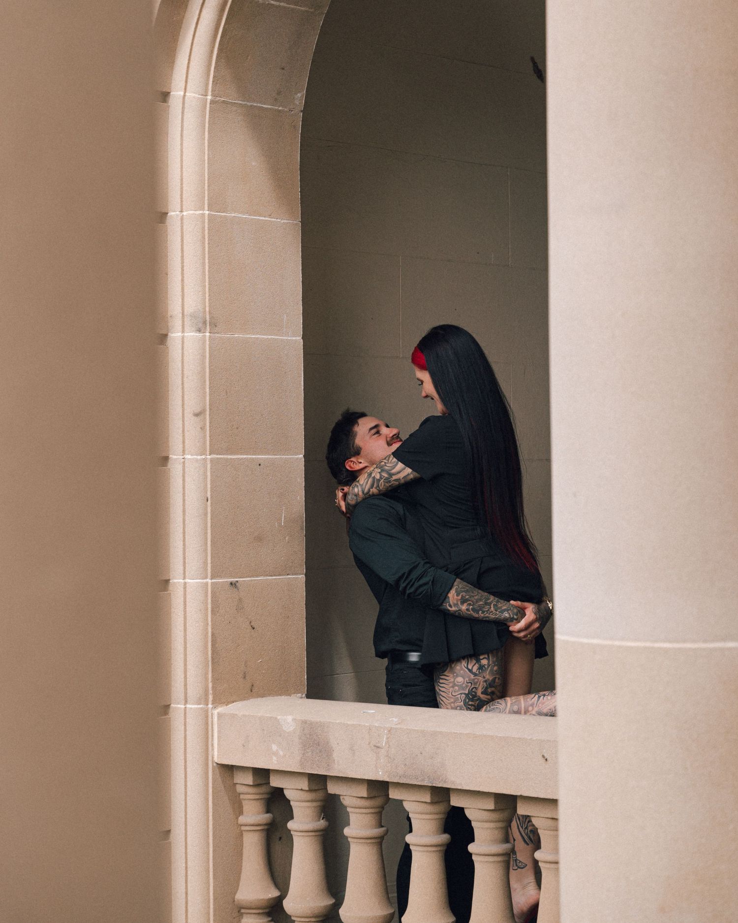 A tattooed man in dark clothing lifts and embraces a long-haired woman with a red hairpiece on a stone balcony beneath an arched doorway.