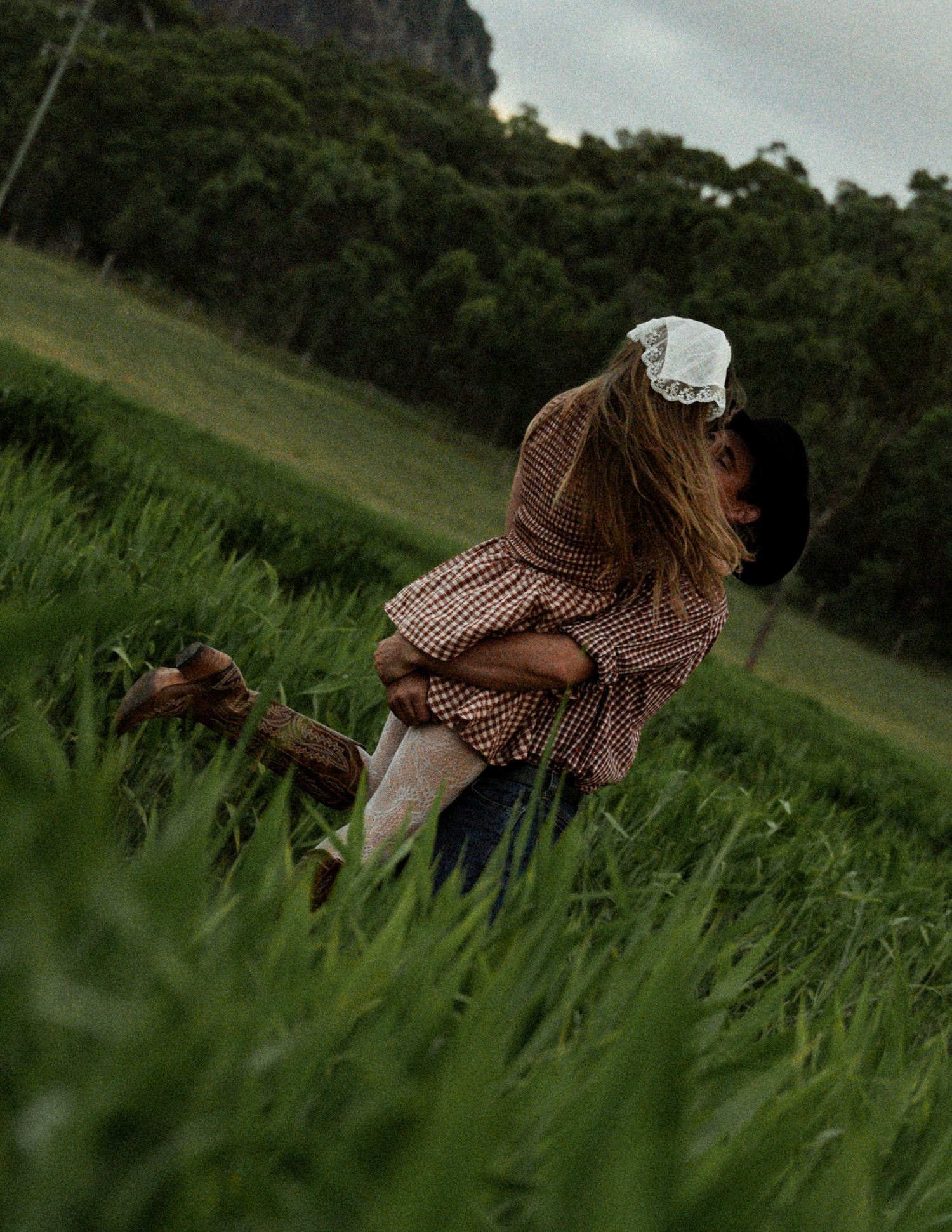 Couple embraces in a romantic lift in a lush green grassy field, the woman wearing a gingham top and white lace headscarf.