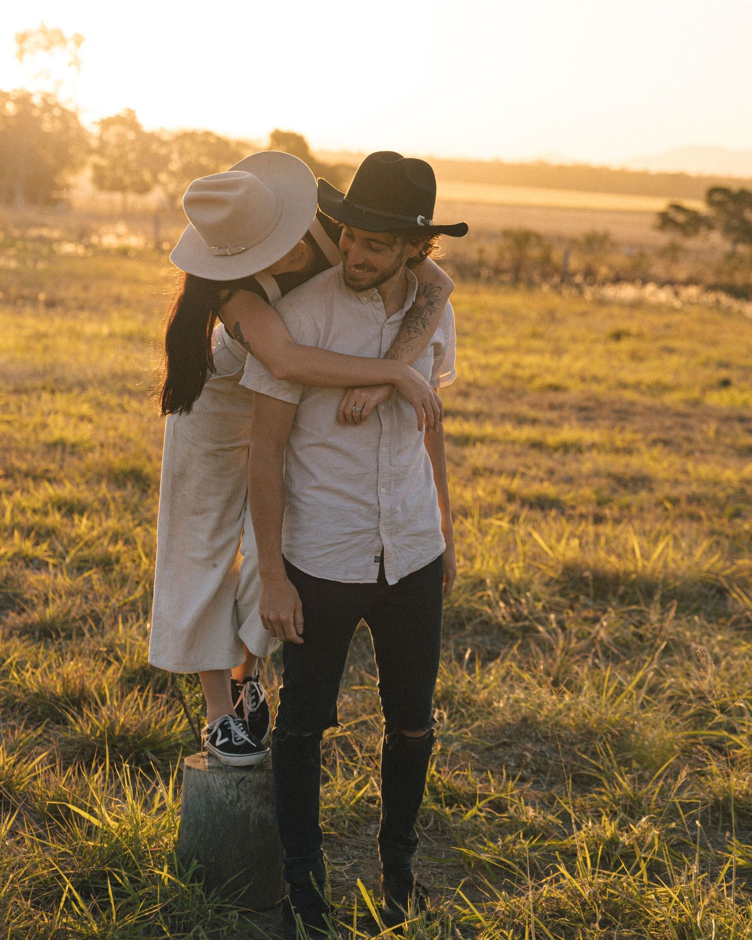 Woman in a wide-brim hat hugs a man in a black cowboy hat from behind in a sunlit golden meadow.