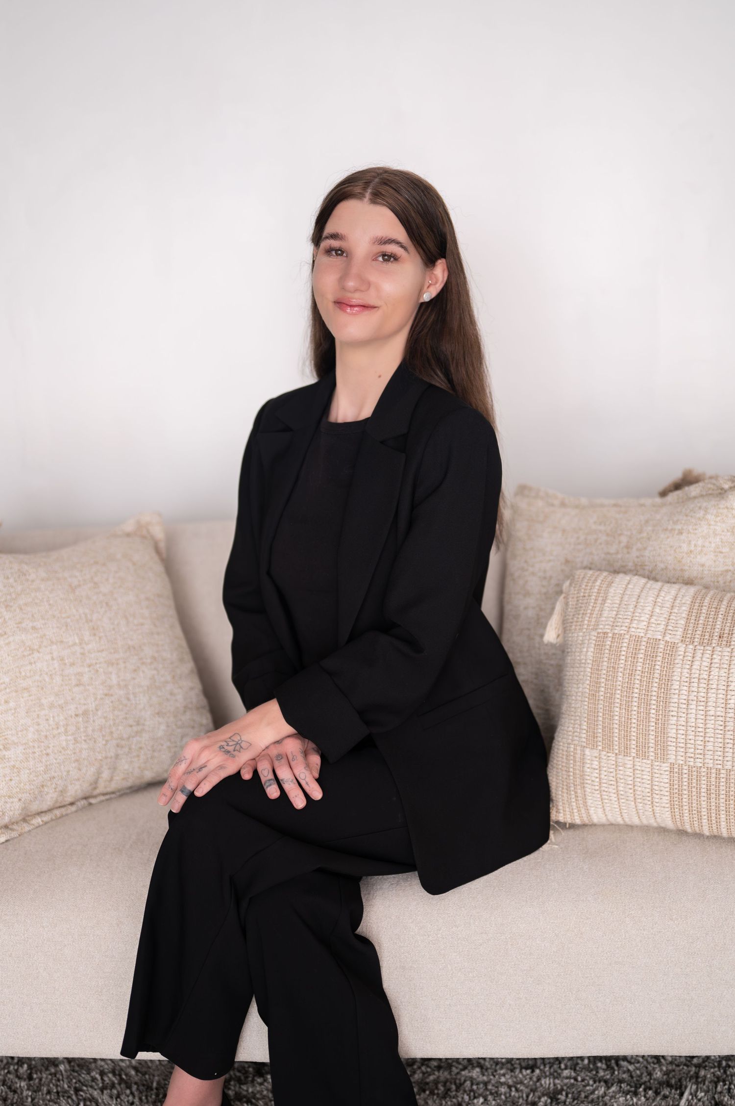 Woman with long brown hair in a black blazer sits on a cream sofa with textured pillows, hands resting on her lap.