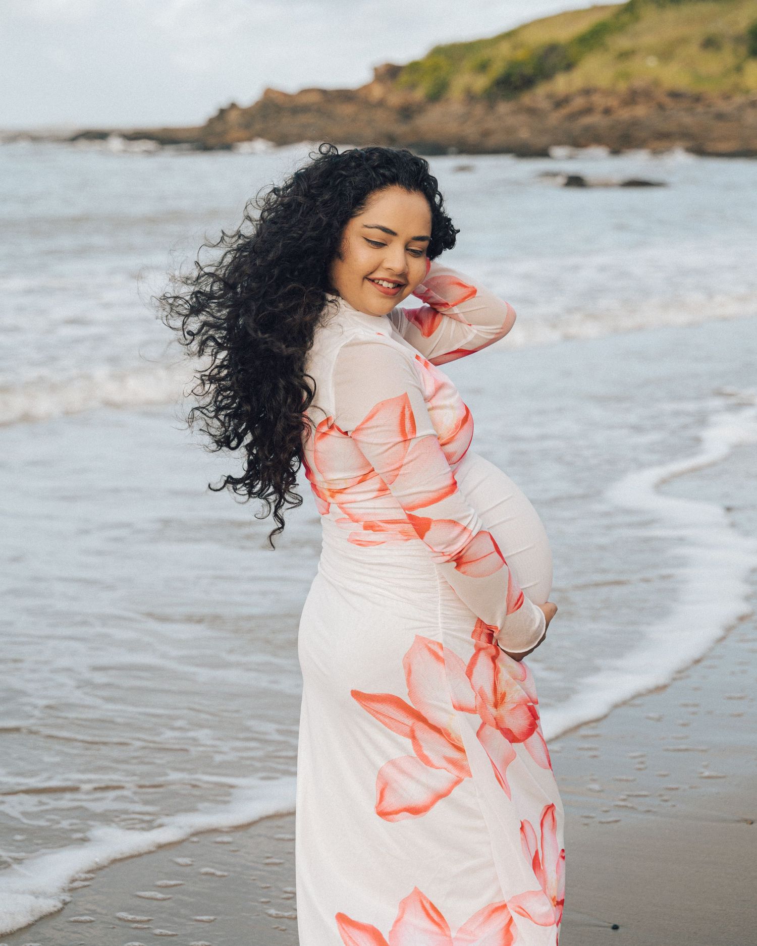 Joyful pregnant woman with curly hair laughs and looks over her shoulder while standing on a beach in a floral maternity gown.