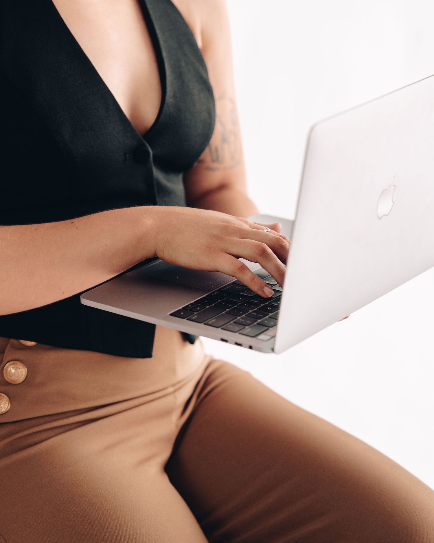 Woman in a black vest and tan trousers typing on a silver laptop held on her lap in a bright studio.