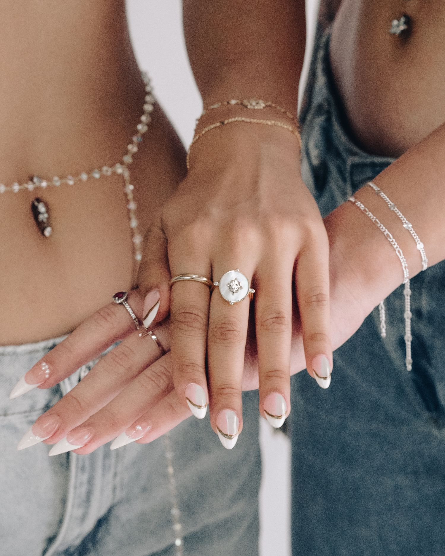 Two people showing off layered gold and silver bracelets, statement rings and belly chain jewelry with long nude manicured nails.
