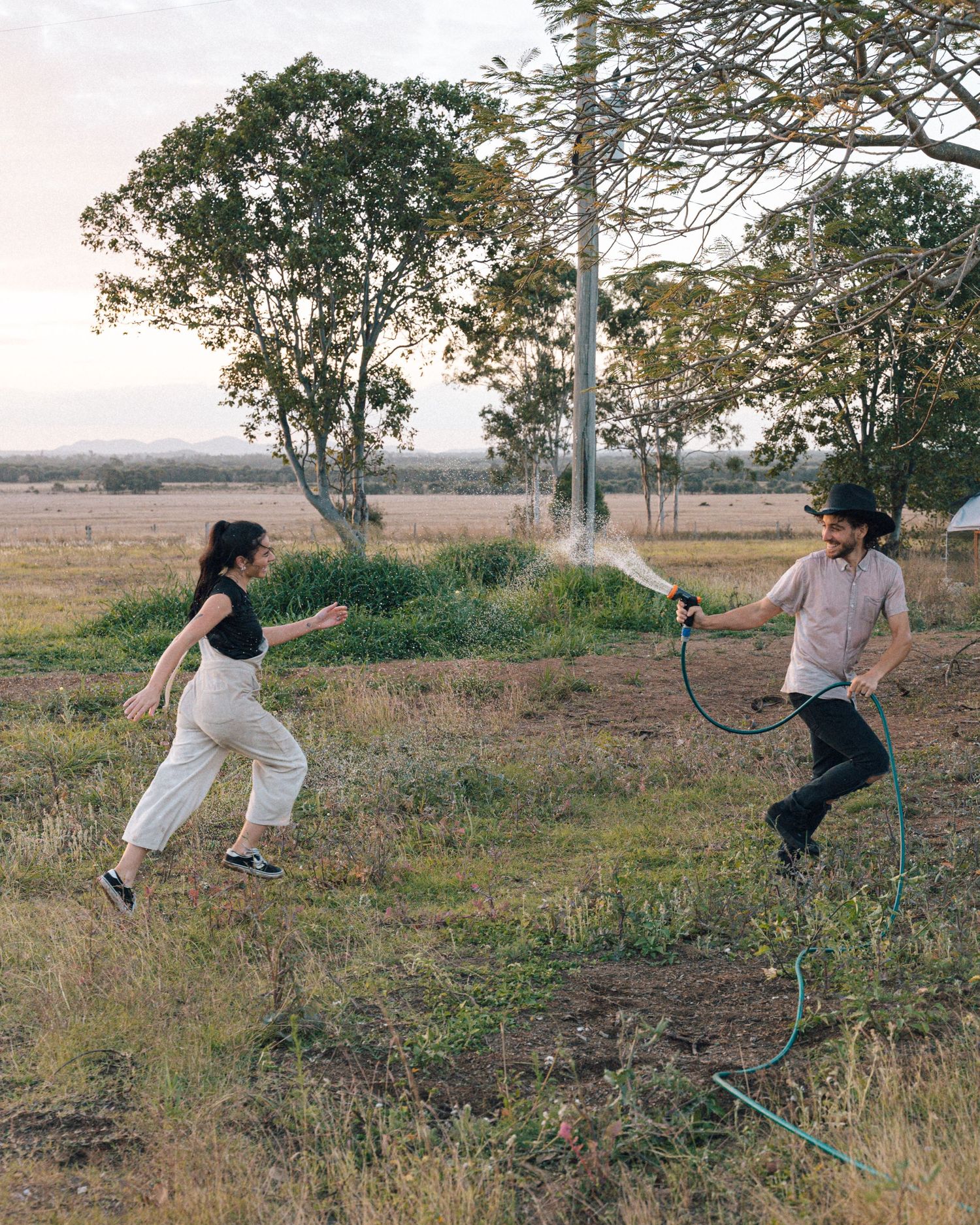 A man in a black hat sprays water from a garden hose as a woman in white overalls runs through a grassy field.