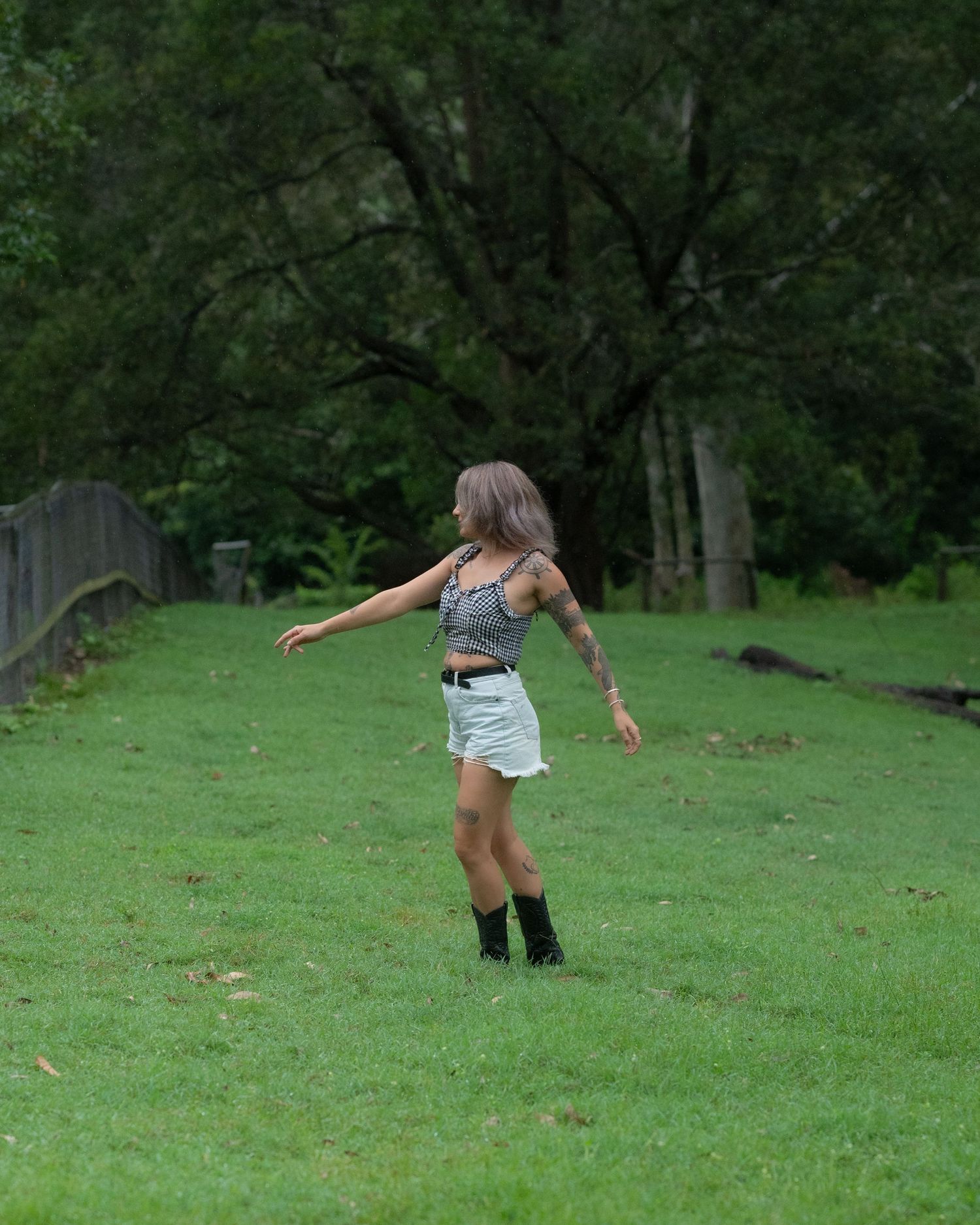 Woman with tattooed arms and black boots twirls with arms outstretched on a grassy hillside surrounded by lush green trees.