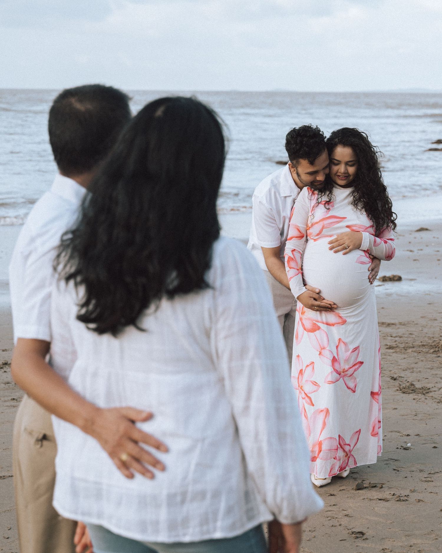 Expectant couple embrace on the beach with her pregnant belly showing while two blurred family members watch in the foreground.