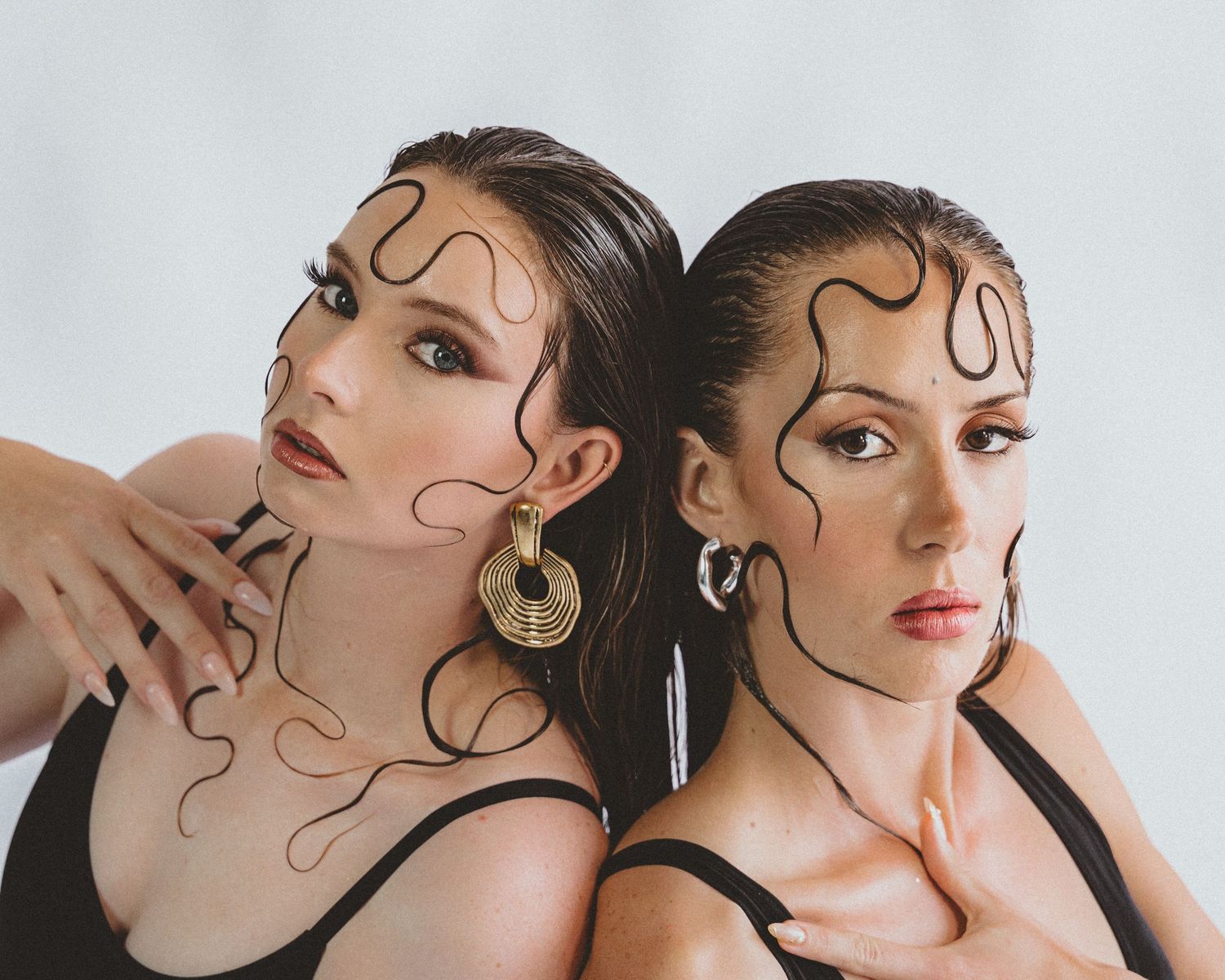 Two women with sleek wet-look hair styled in dramatic swirling designs pose back to back in black tops and statement gold earrings.
