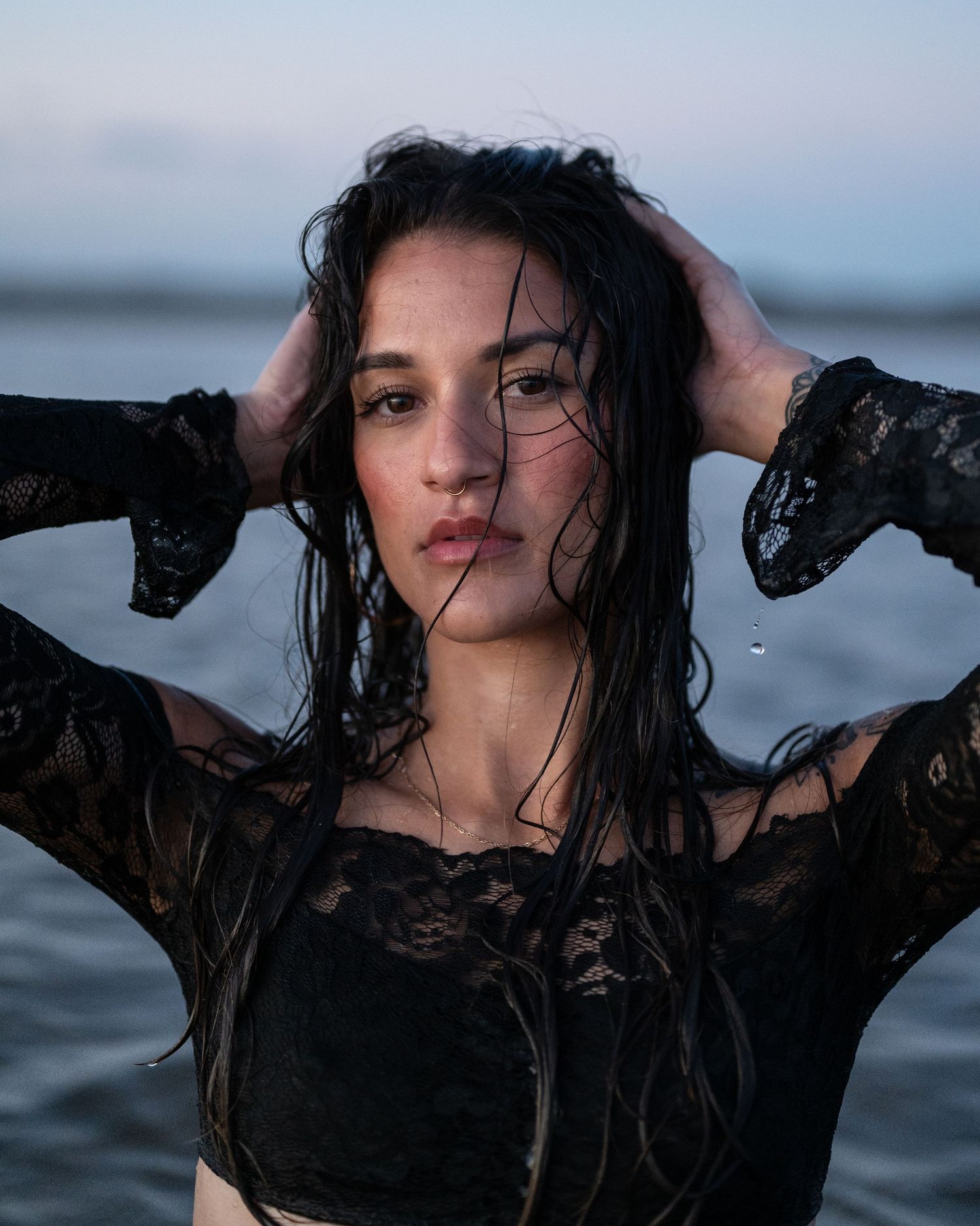 Portrait of a woman with wet dark hair and black lace sleeves raising her hands to her head while standing in the water at dusk.