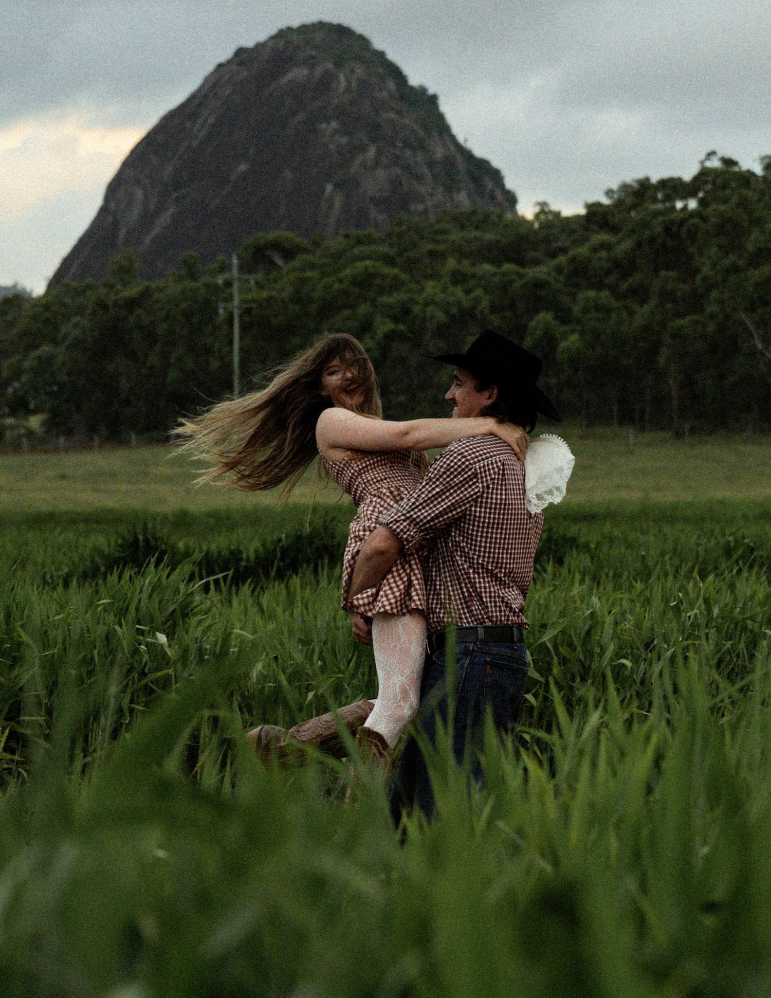 Man in a black hat lifting a woman with long flowing hair in a green field beneath a rugged mountain and moody sky.