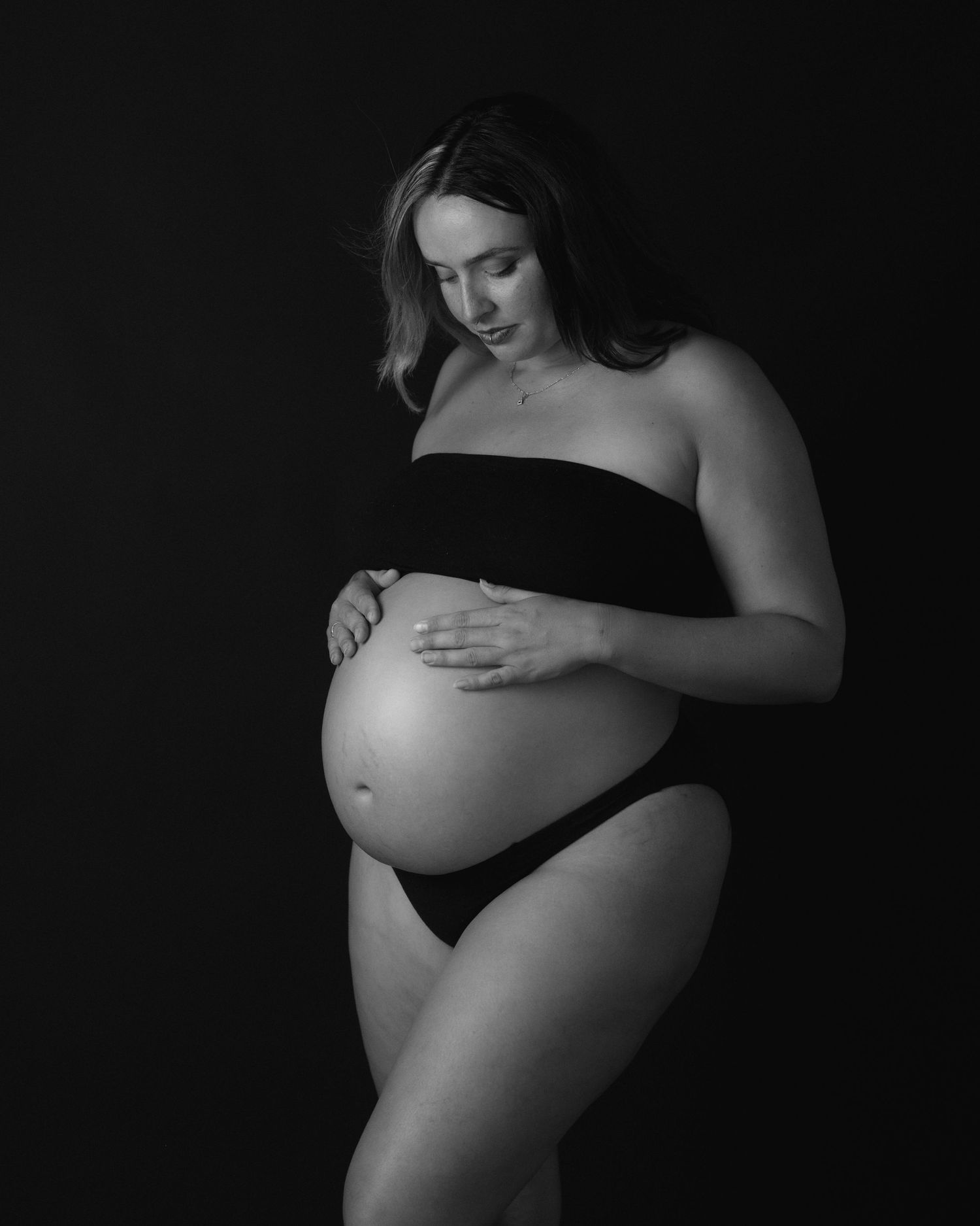 Black and white maternity portrait of a pregnant woman in dark two-piece styling looking down at her belly.