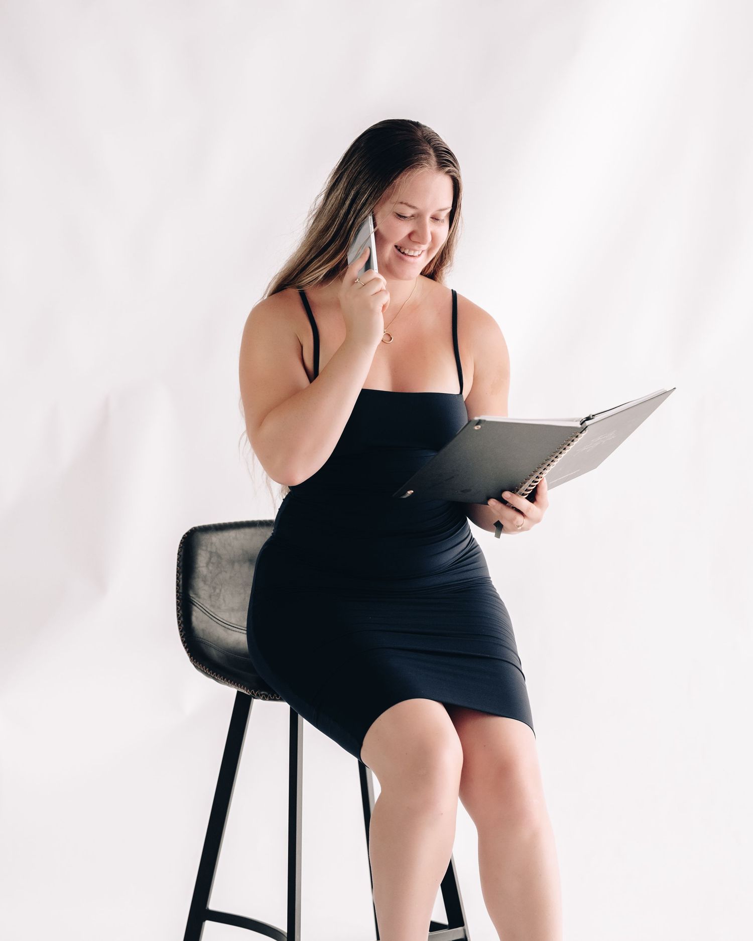 A smiling woman in a black dress sits on a bar stool, talking on her phone while holding an open folder.