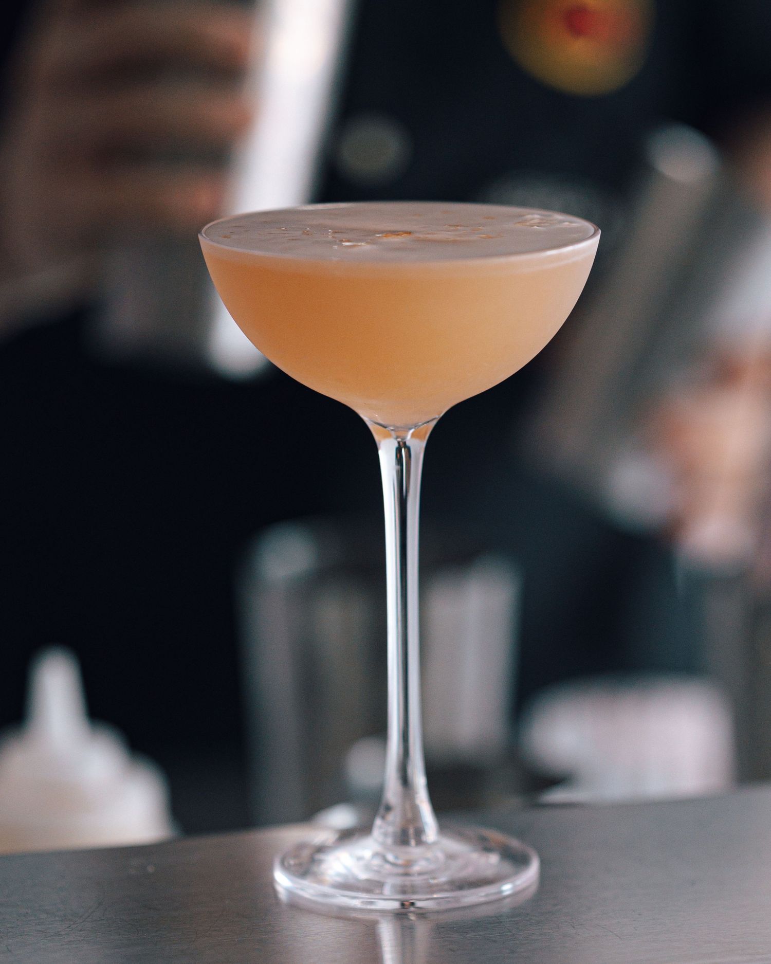 Pink frothy cocktail served in a coupe glass on a stainless steel bar with a bartender blurred in the background.