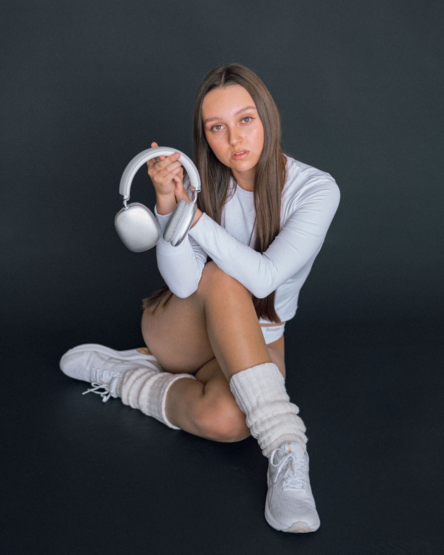 Woman sits on the floor holding white over-ear headphones, dressed in a white top, leg warmers and sneakers against a dark backdrop.