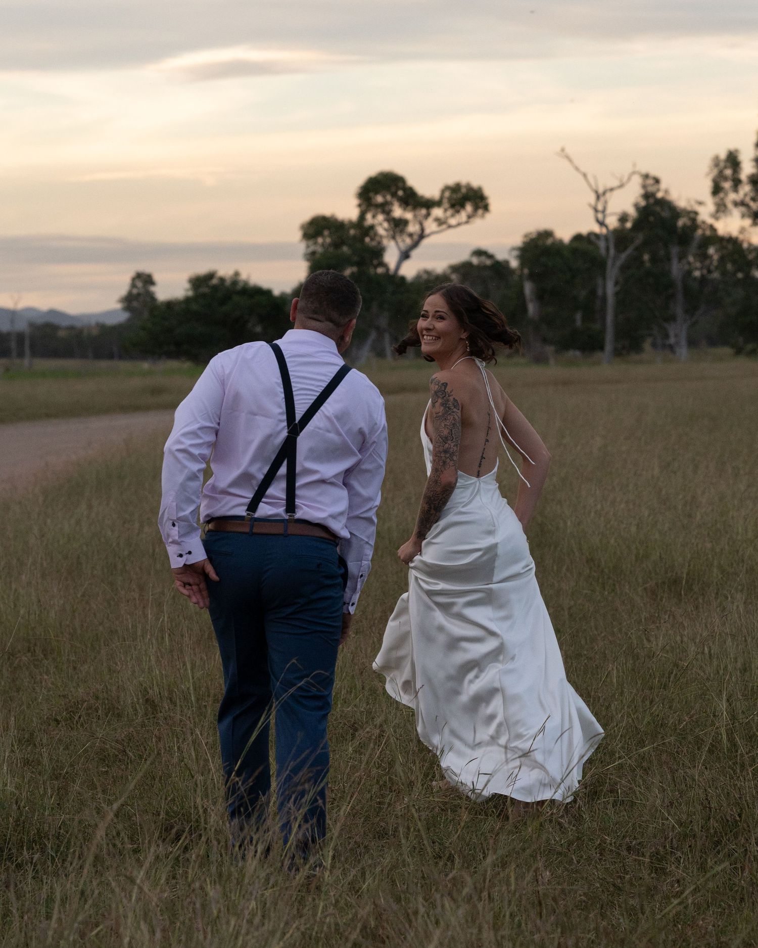 Bride in a white gown smiles over her shoulder as she walks with the groom through a golden field at sunset.