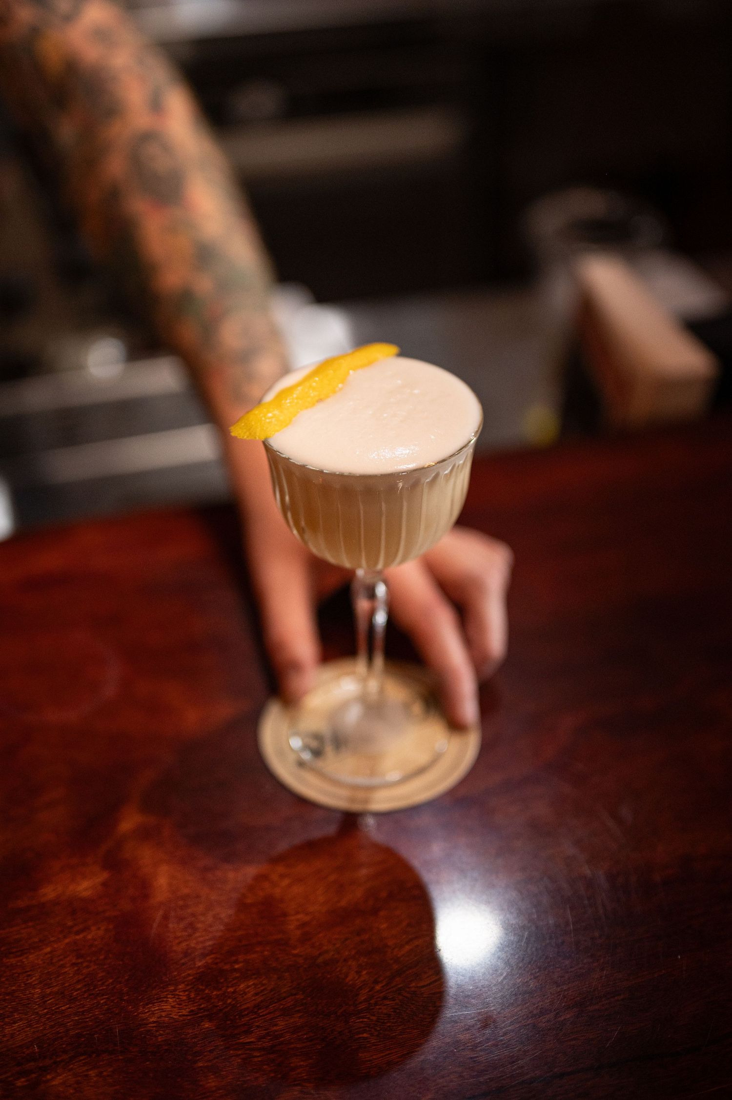 Tattooed hand presents a creamy white cocktail garnished with a lemon peel in a ribbed coupe glass on a glossy wooden bar top.