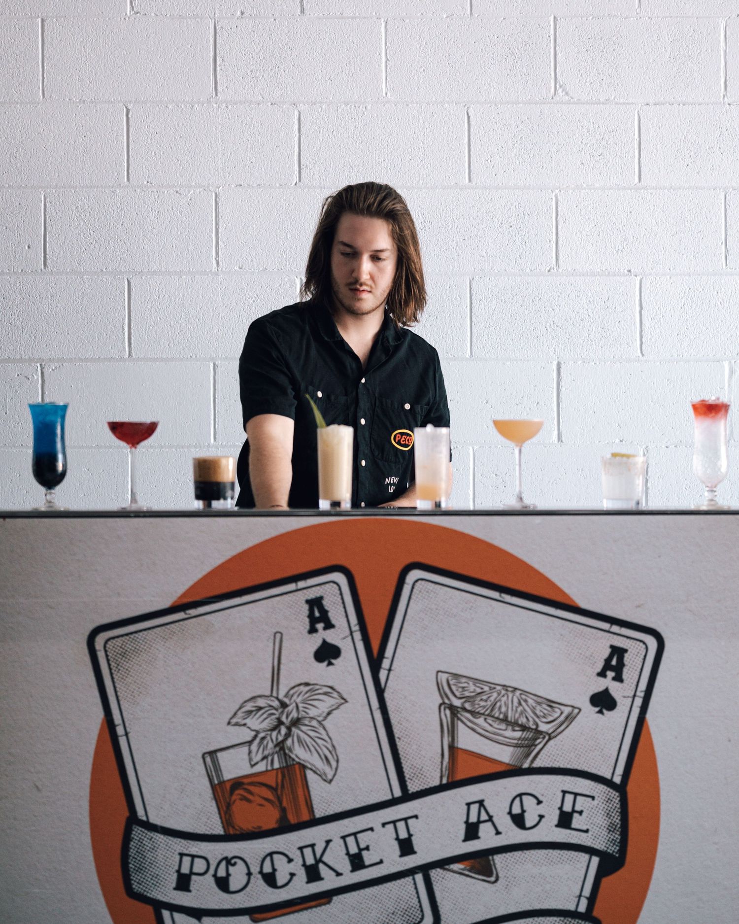 Bartender in black shirt stands behind a Pocket Ace Bartending bar lined with colorful cocktails in assorted glasses.