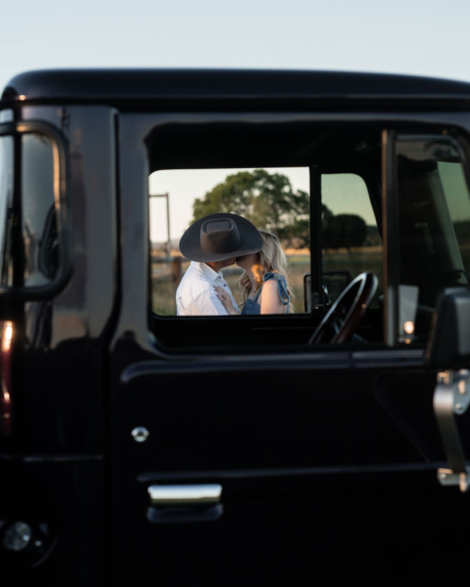 A couple in cowboy hats shares a kiss, framed through the open window of a black vintage 4x4 truck.
