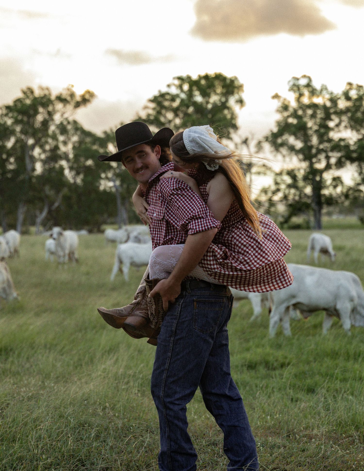 Cowboy in a black hat carries a woman in a gingham dress and boots piggyback style across a pasture with white cattle grazing nearby.