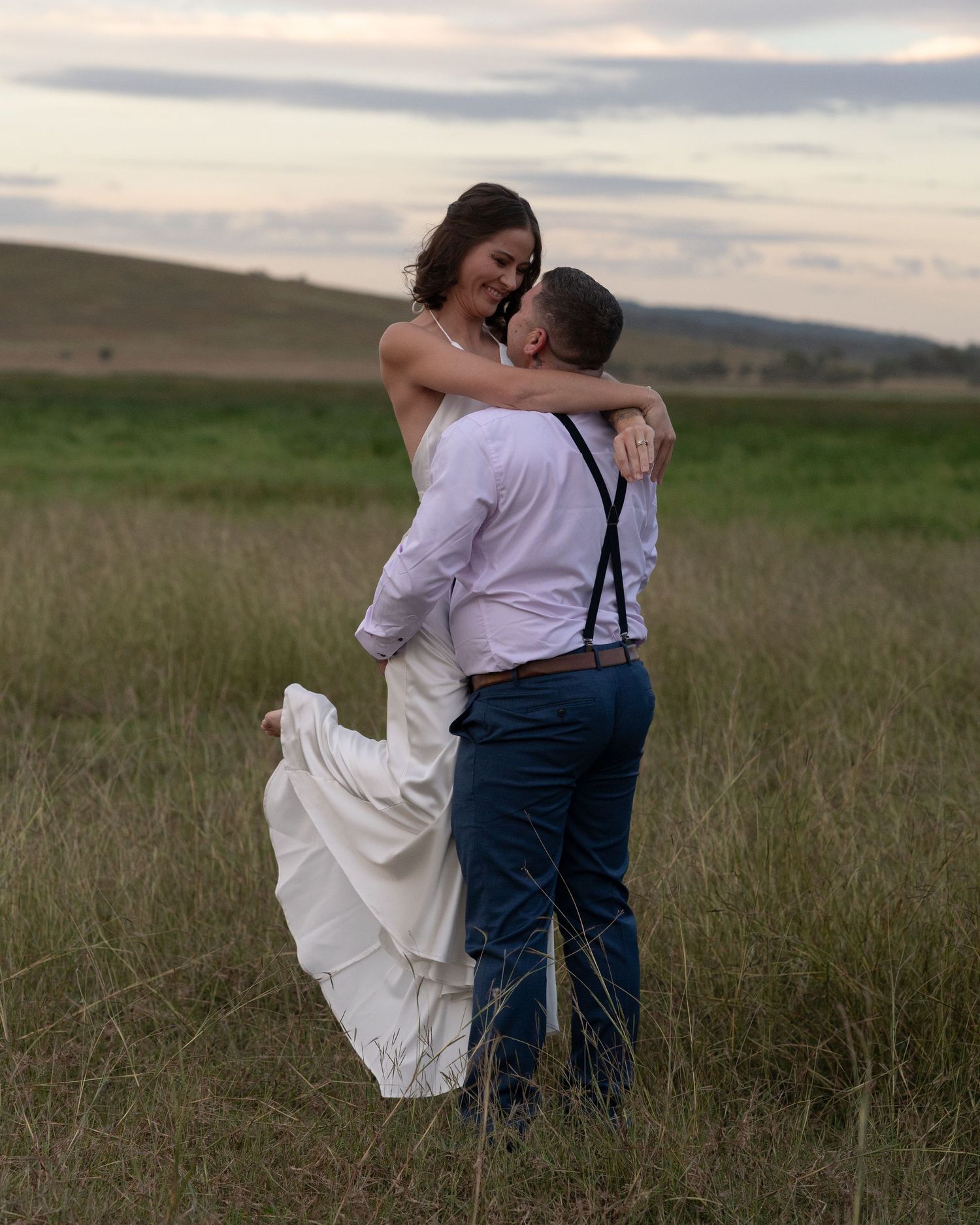 Groom in suspenders and blue trousers lifts his bride in a flowing white gown amid tall grass at twilight.