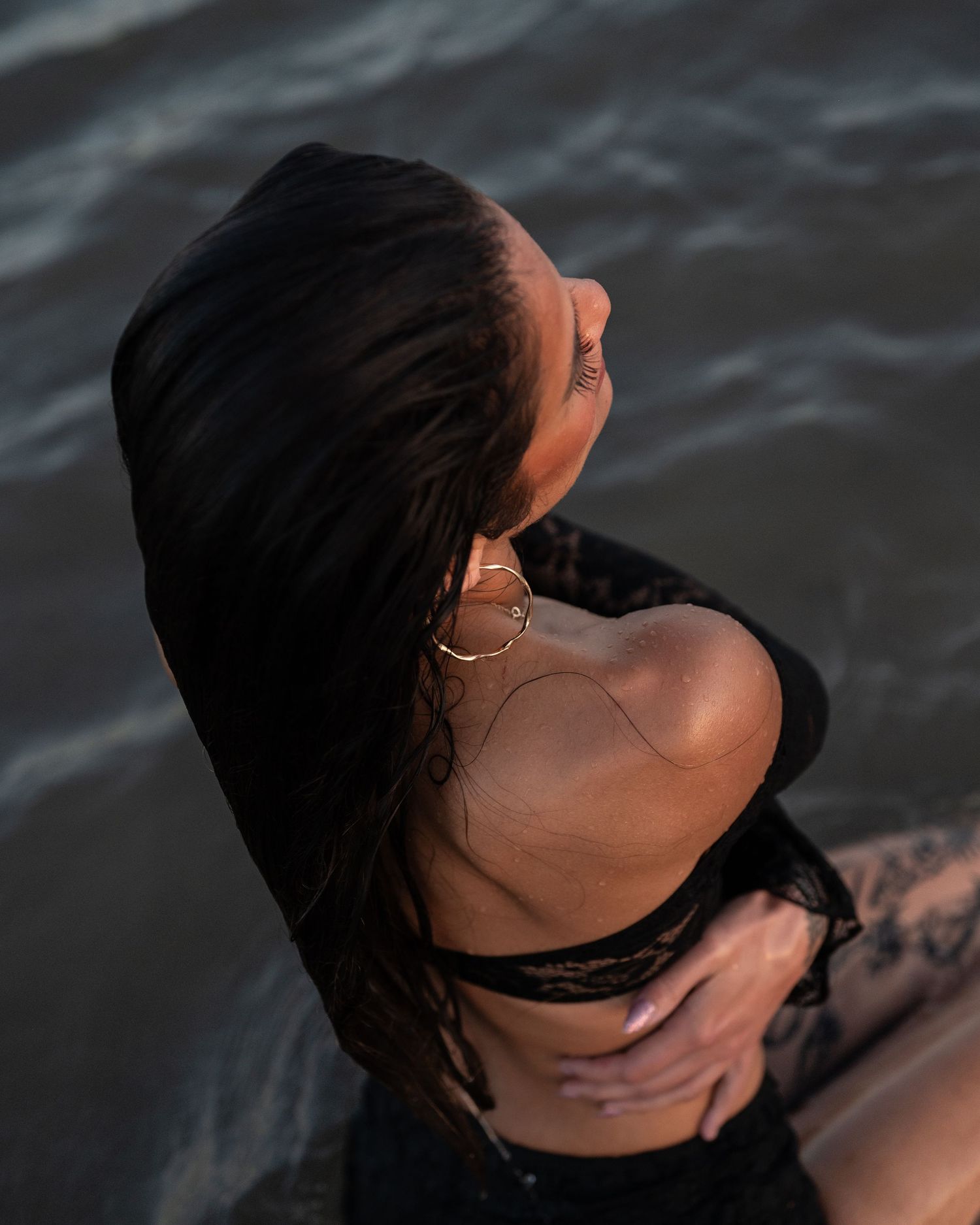 Overhead view of a woman with long wet dark hair, gold hoop earrings and black lace outfit sitting beside dark rippling water.