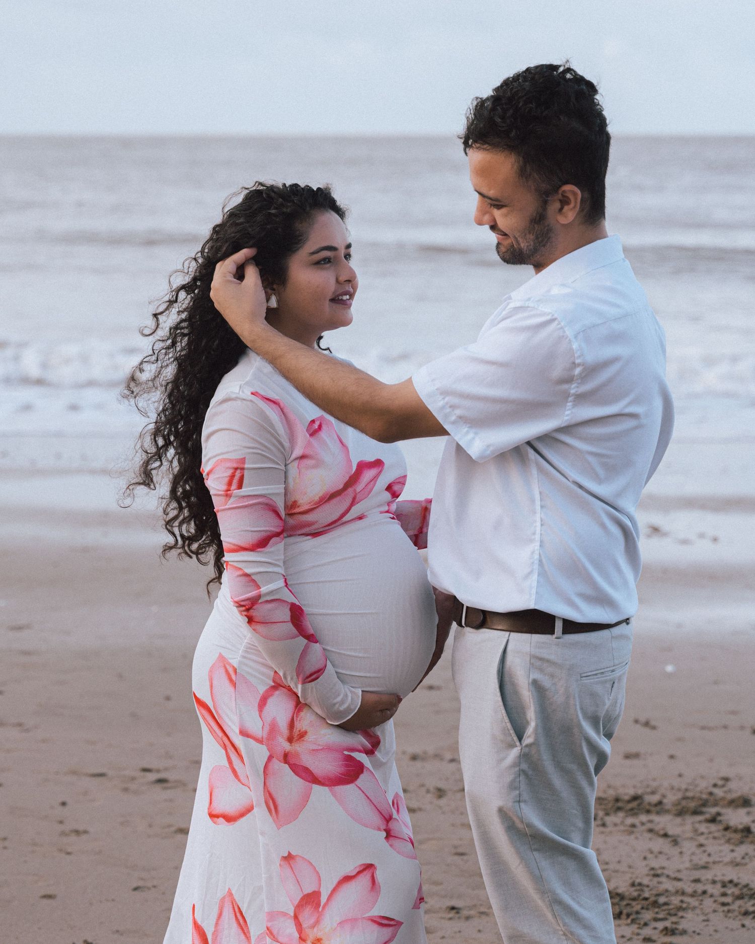 Expectant father smiles and touches his partner's hair on the beach as she wears a pink floral maternity dress.