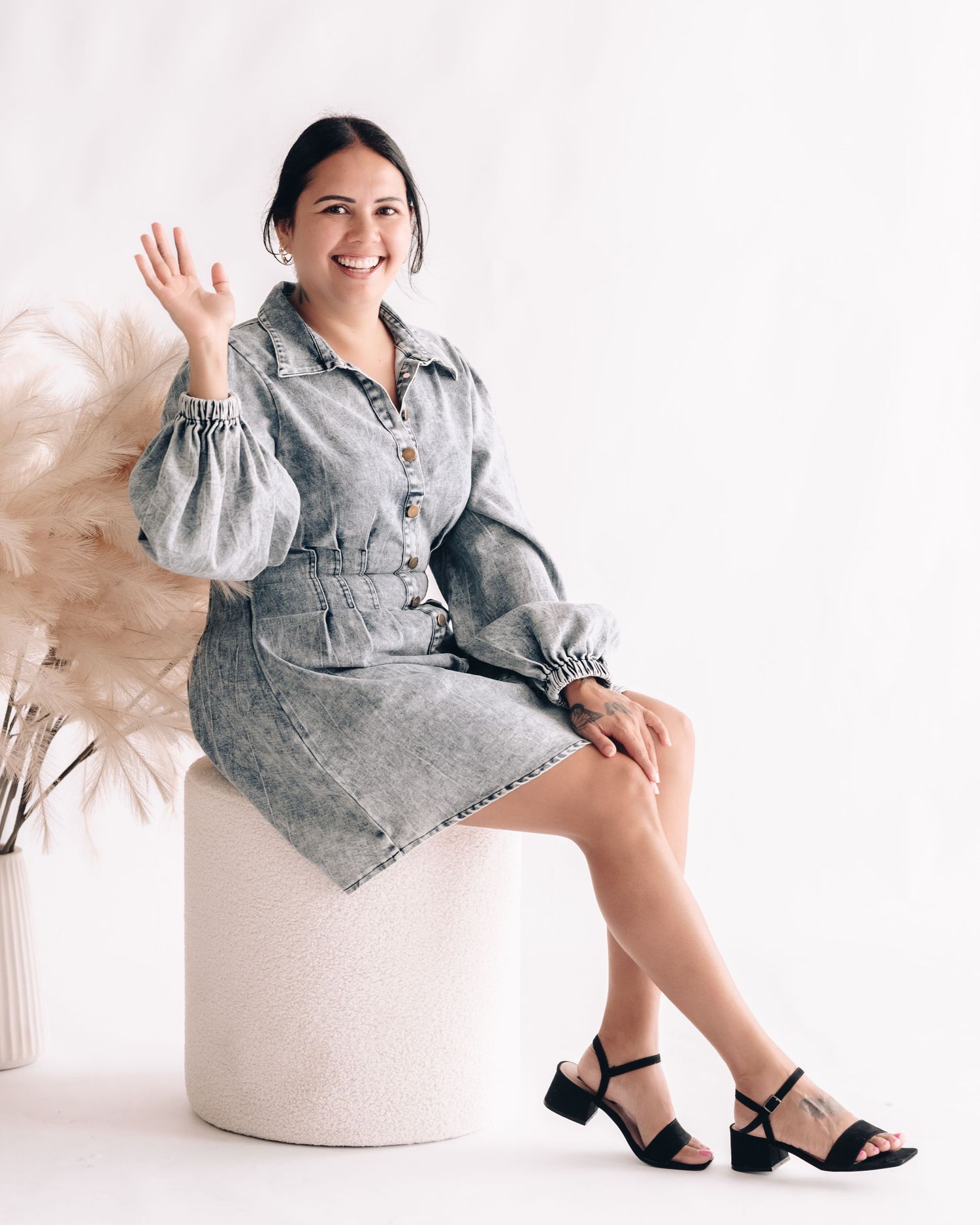 Smiling woman in a gray denim dress waves while seated on a cream stool beside dried pampas grass.