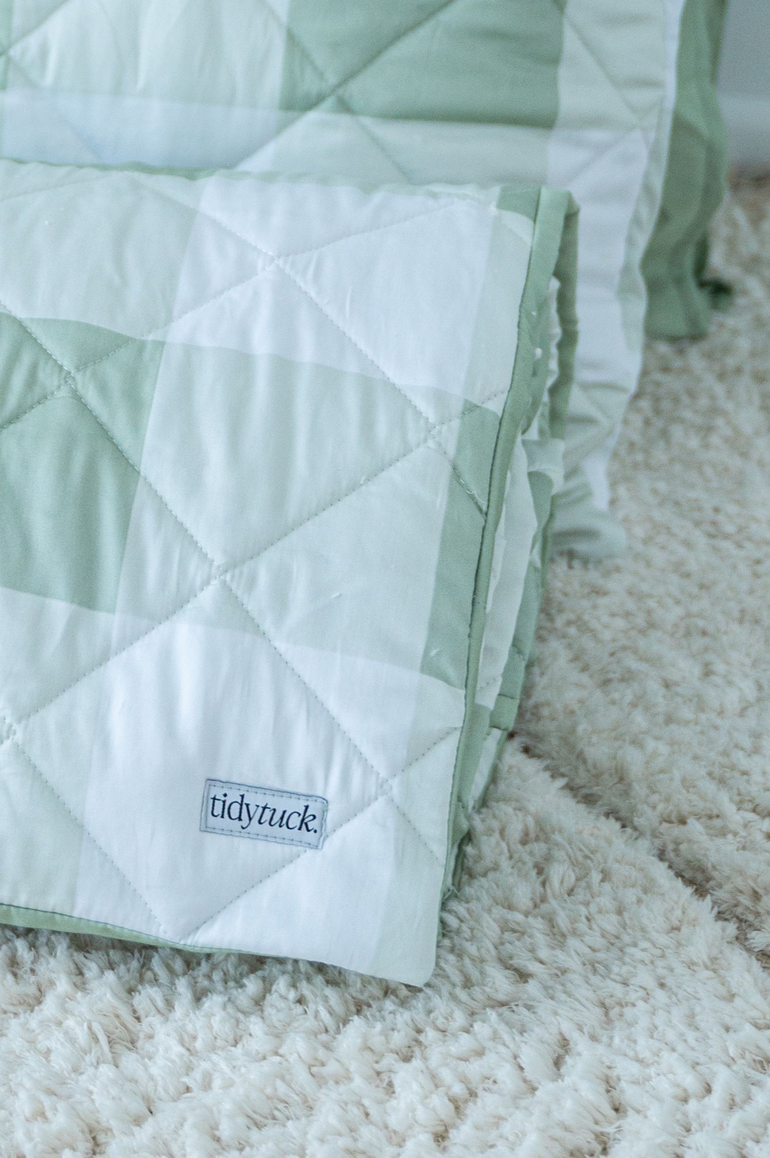 Folded sage green and white gingham quilt with a Tidy Tuck label resting on a soft, fluffy cream shag rug.