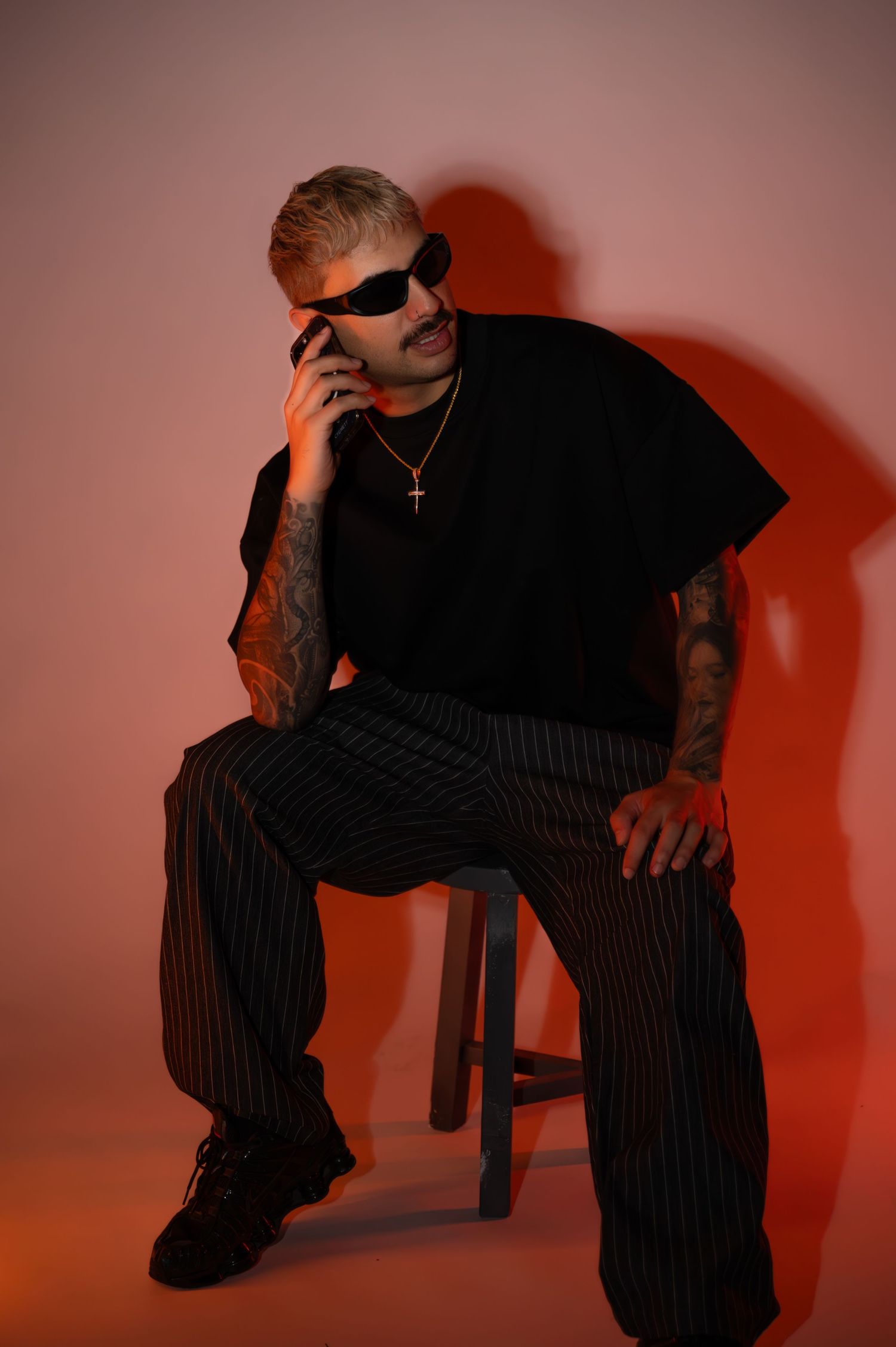 Man in dark sunglasses, black tee and striped trousers sits on a wooden stool talking on his phone against a warm red backdrop.