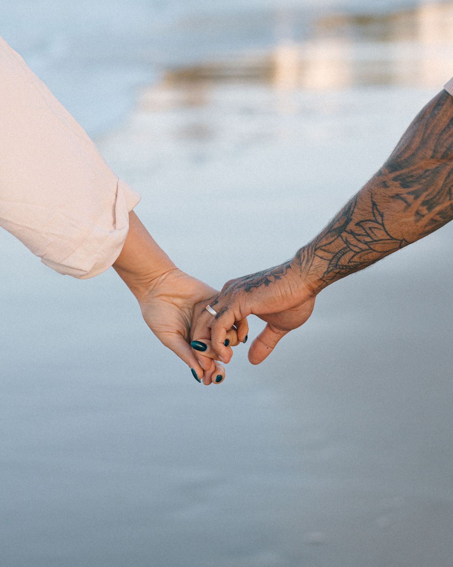 Close-up of two hands held together on a beach, one with a detailed floral tattoo sleeve, with soft shoreline in the background.